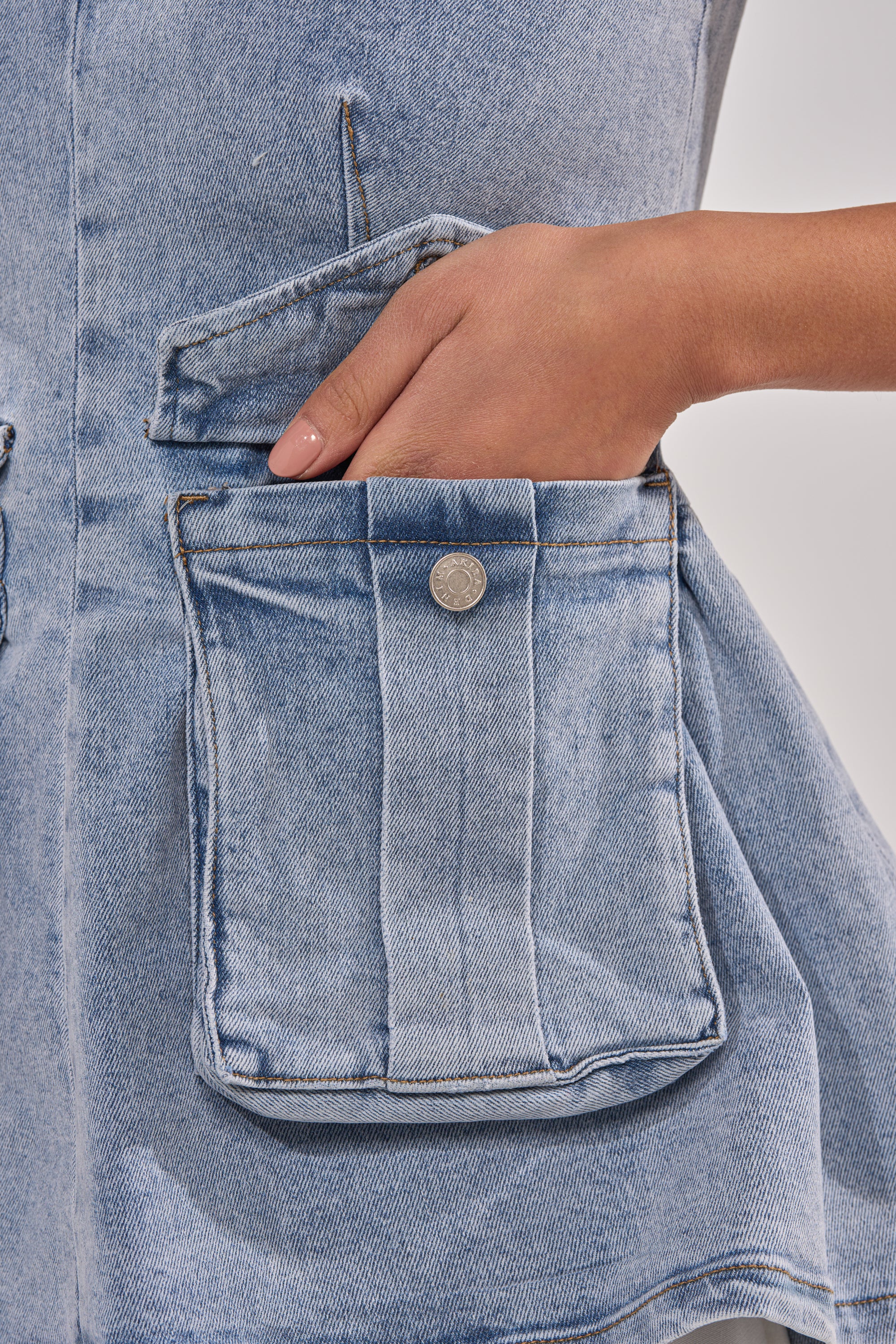 A close-up shows a hand sliding into the button-flap pocket of the TURN AROUND DENIM BLOUSE, highlighting the seam and texture of this light blue denim peplum top for a chic, functional utility style. 6