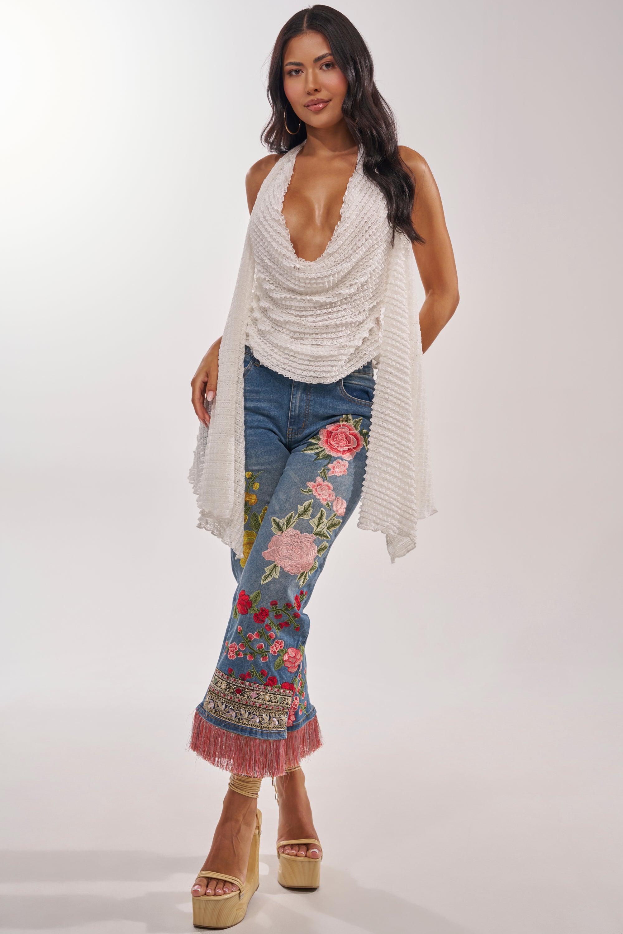 A woman stands wearing the TAKE ME TO THE TROPICS COWL NECK BLOUSE, blue jeans with colorful floral embroidery and pink fringe at the hem, and beige platform sandals against a plain light background. 5