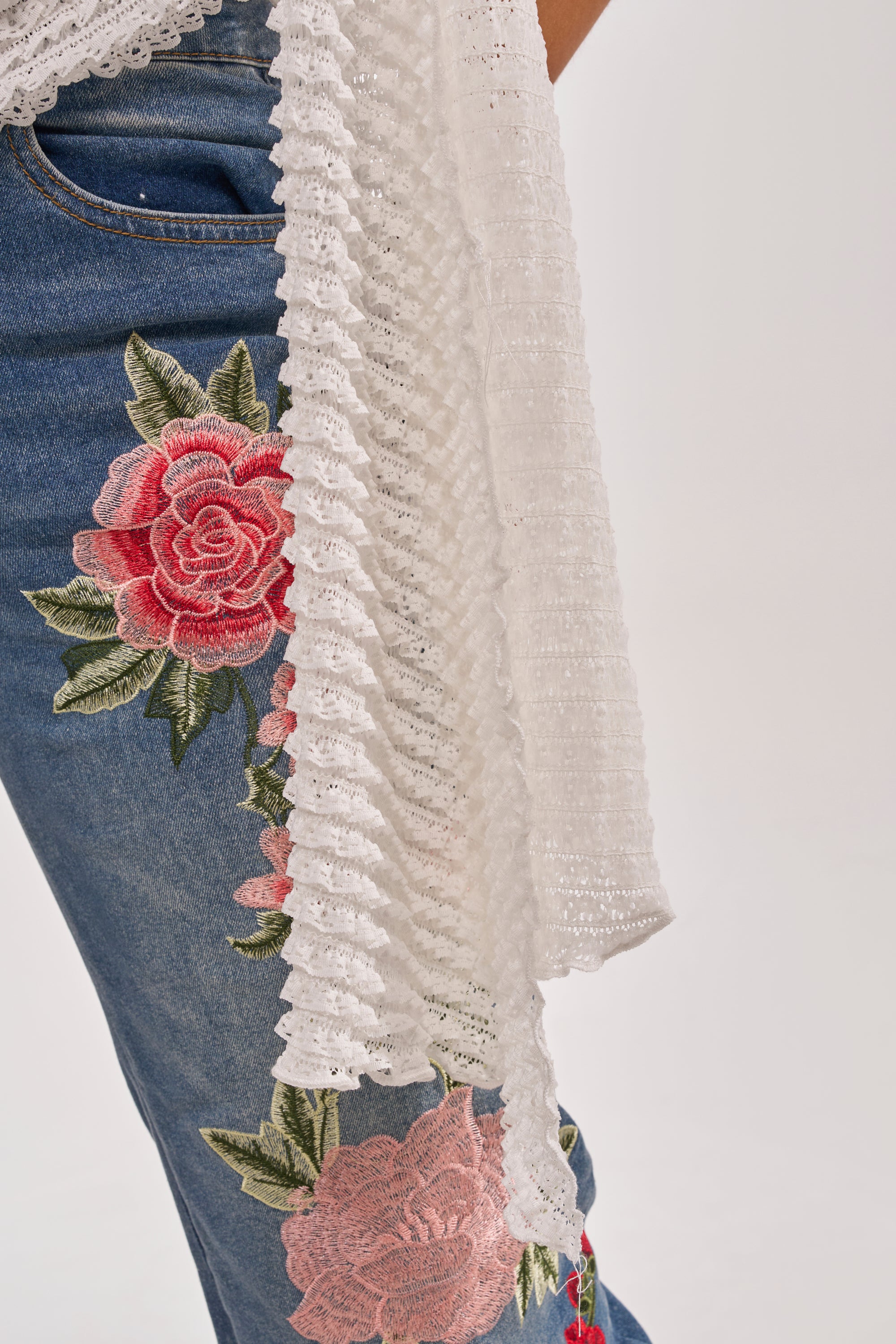 A person wears the TAKE ME TO THE TROPICS COWL NECK BLOUSE, styled with blue jeans featuring large pink embroidered flowers and a draped white lace fabric, set against a plain light background. 8