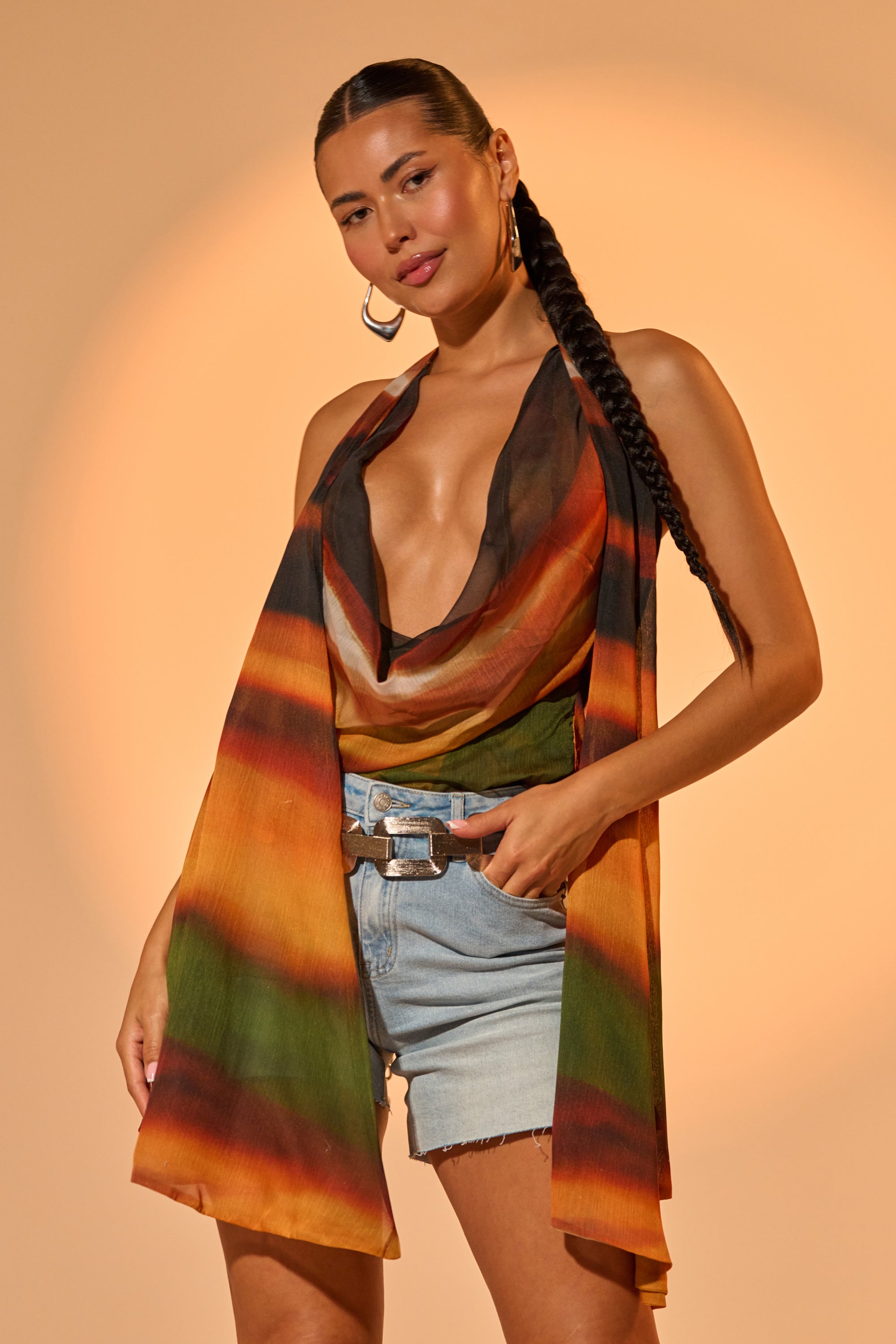A woman with long braided hair models the SELENA DEEP COWL HALTER TOP, a colorful halter featuring a draped cowl neckline, paired with light blue denim shorts. She poses confidently with one hand on her hip against a warm beige backdrop. 0