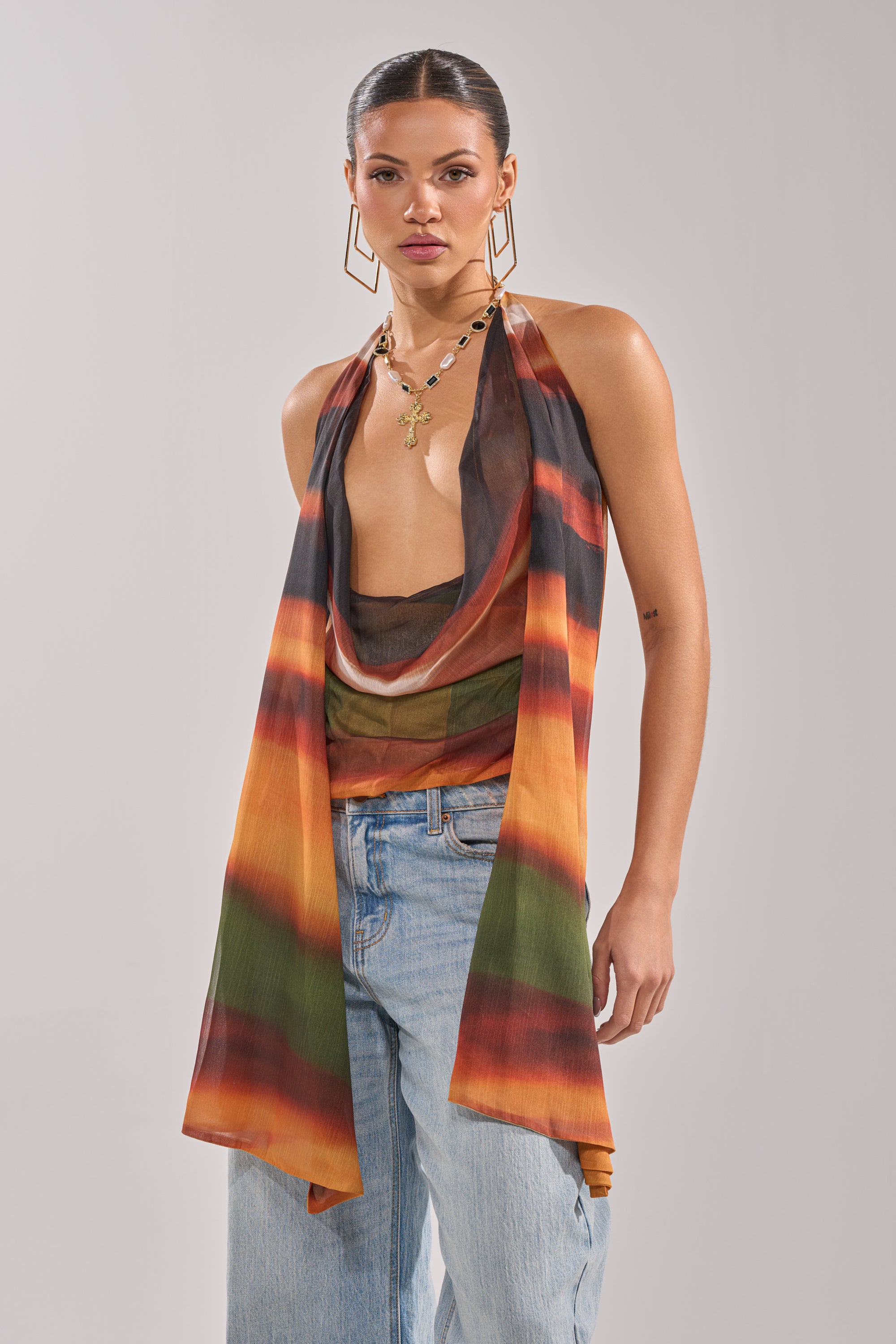 A woman stands against a plain light background wearing the SELENA DEEP COWL HALTER TOP in a multicolor, sleeveless style with an open front, paired with light blue jeans, layered necklaces, and large square hoop earrings. 2