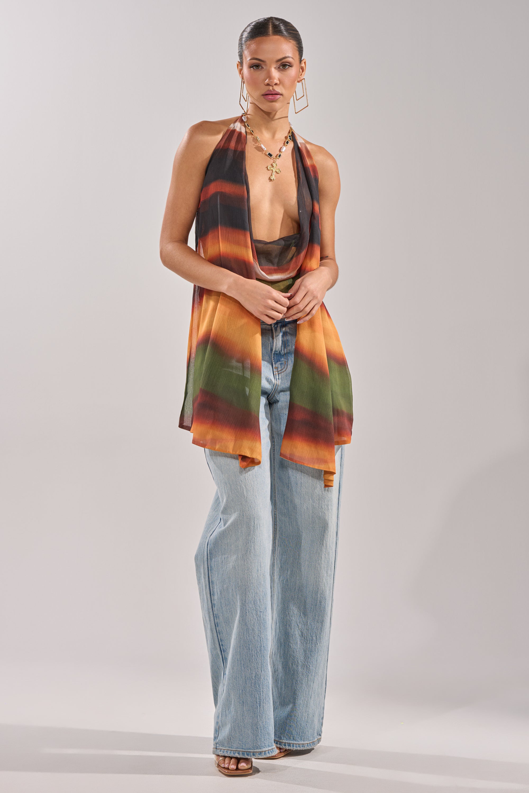 A woman wears the SELENA DEEP COWL HALTER TOP—an open-back, sleeveless halter with orange, black, green, and brown stripes—paired with light blue wide-leg jeans, gold jewelry, and black sandals against a plain background. 7
