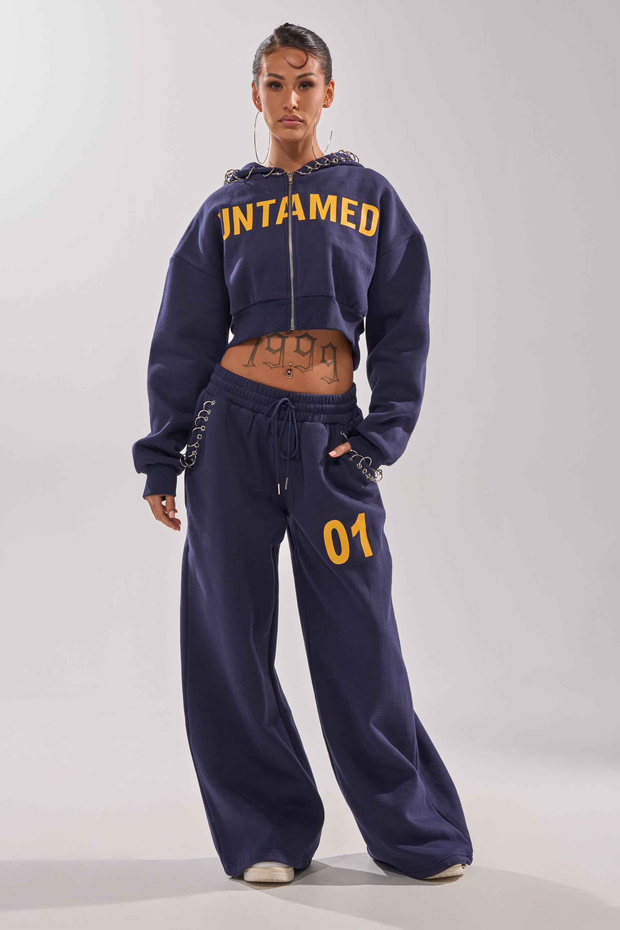 A woman faces forward in UNTAMED METAL RING JOGGERS—navy, wide-leg, high-rise with "01" on one leg—paired with a matching cropped hoodie featuring "UNTAMED" in yellow, hoop earrings, white sneakers, and visible tattoos. 0