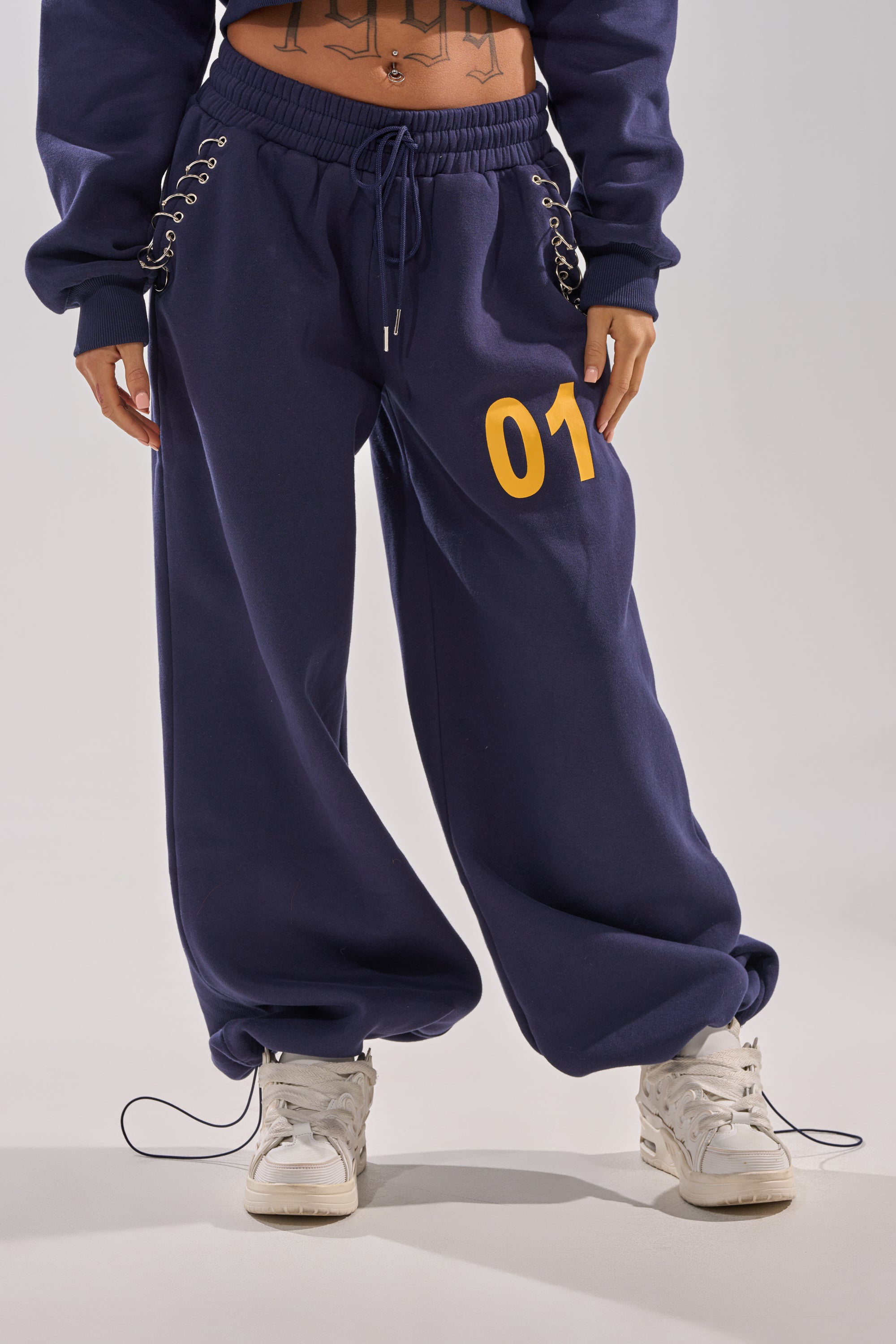 A person wears UNTAMED METAL RING JOGGER—navy blue fleece sweatpants featuring lace-up hips, a yellow "01" on one thigh, and a high-rise elastic waistband—paired with white sneakers. A torso tattoo is visible. 9