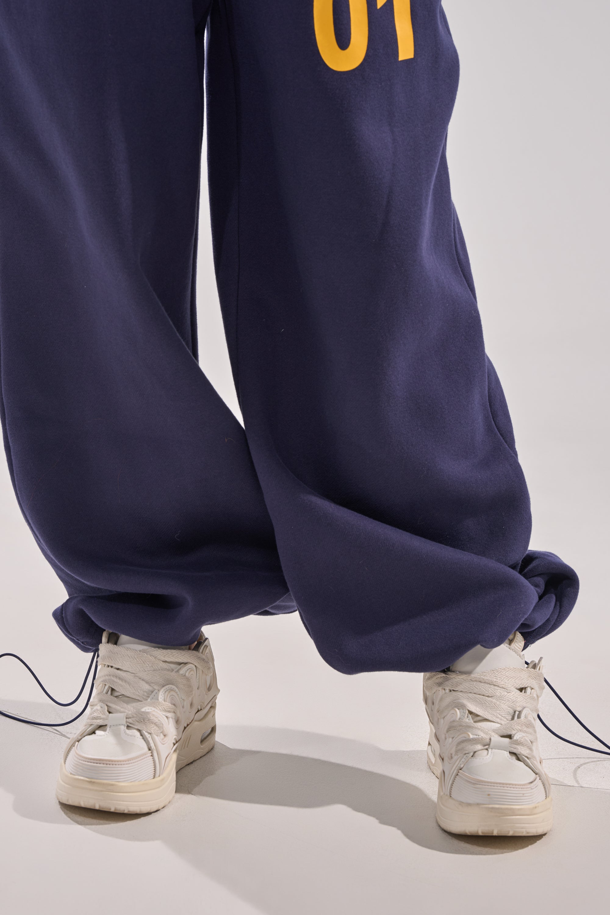 Model wears UNTAMED METAL RING JOGGER—white, loose-fitting sweatpants featuring yellow numbers and a high-rise elastic waistband—styled with white sneakers, standing with feet crossed on a light background. 10
