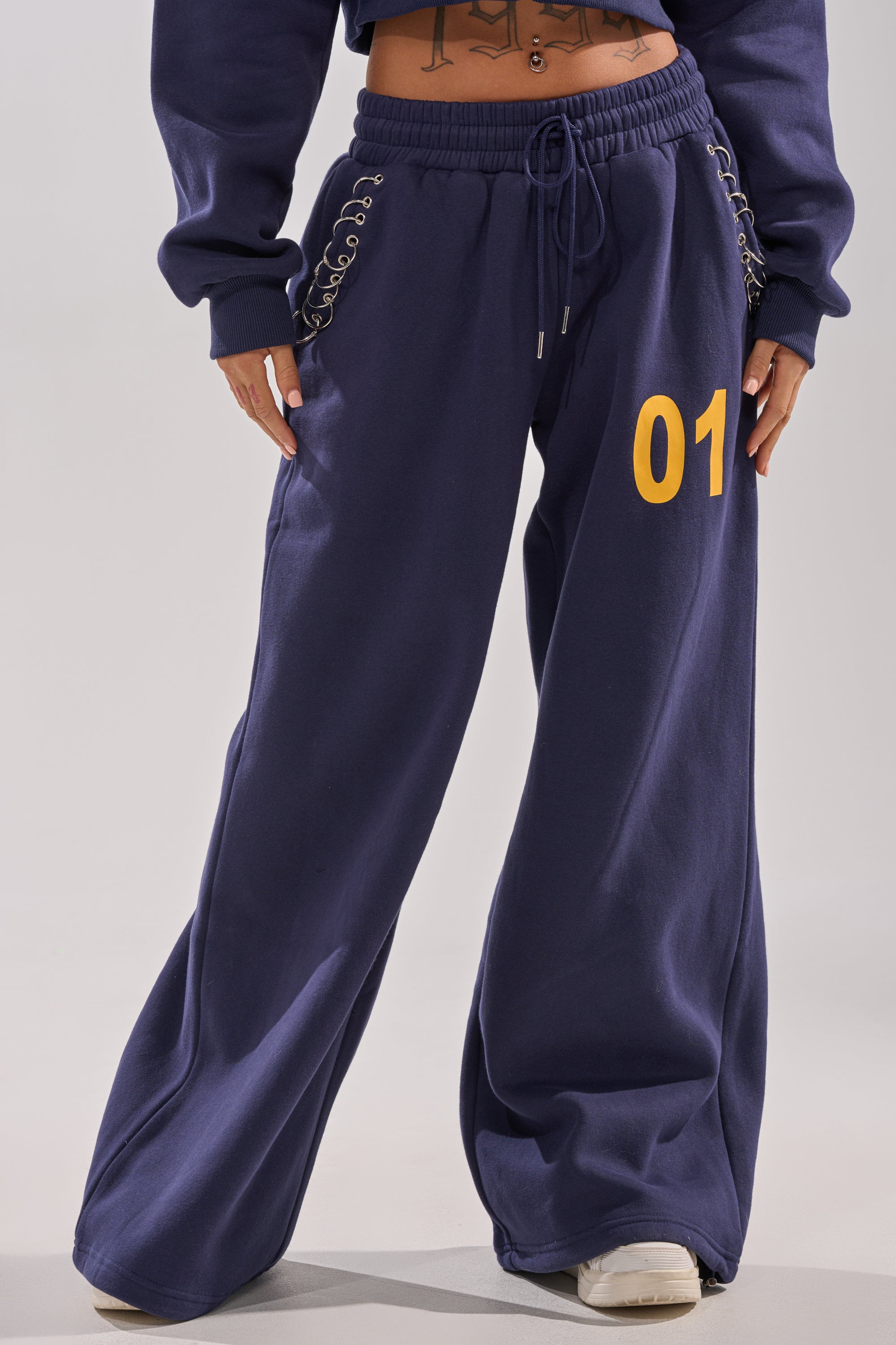 Someone is wearing the UNTAMED METAL RING JOGGER, a pair of high-rise, wide-leg navy sweatpants with a yellow "01" on the left thigh, lace-up hip details, paired with white sneakers. Only their lower half is visible. 1