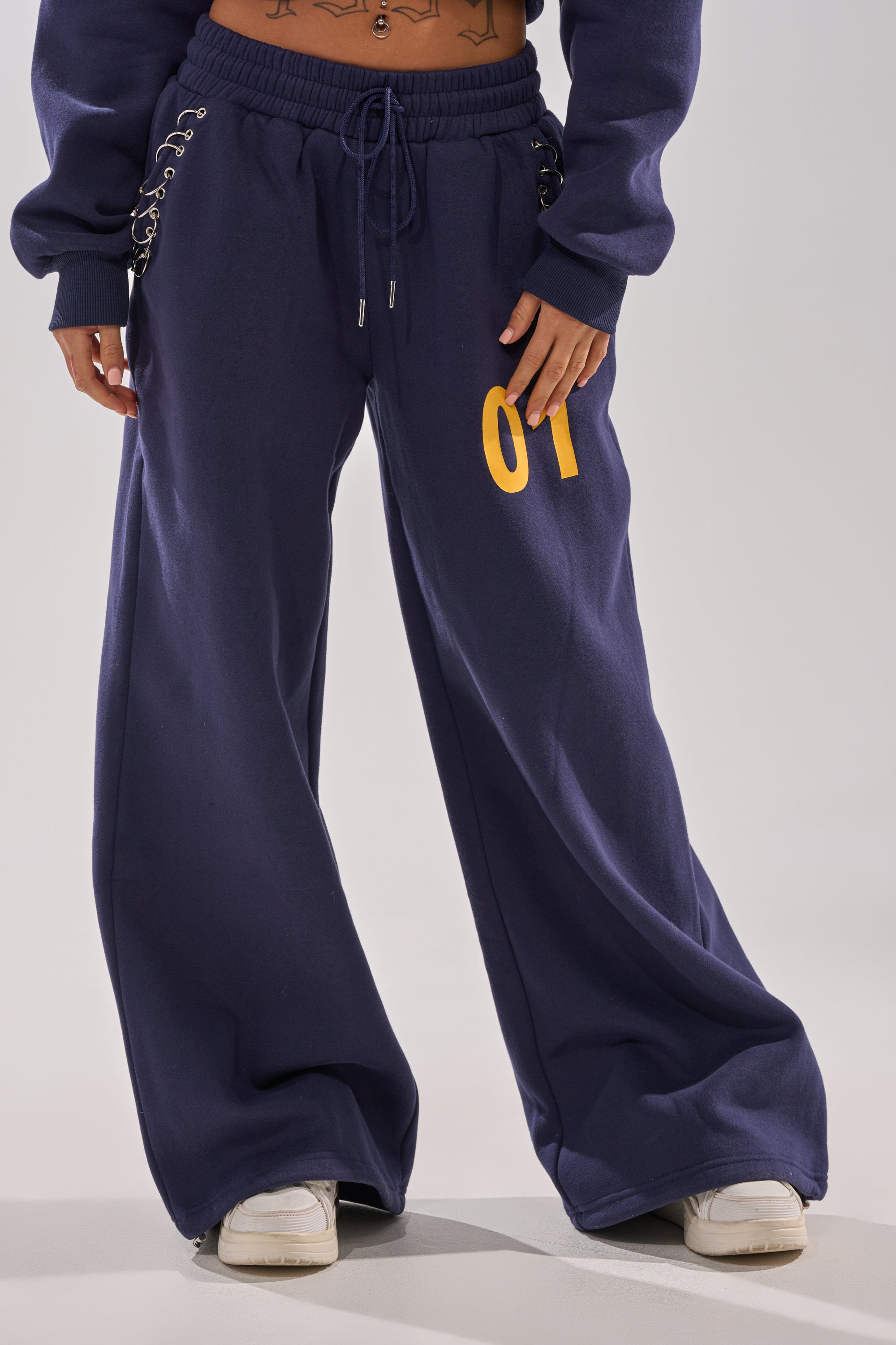 A person wearing UNTAMED METAL RING JOGGER sweatpants in navy blue with a yellow "01" on the left thigh, paired with a matching sweatshirt and white sneakers. Only the lower half of the body is visible. 2