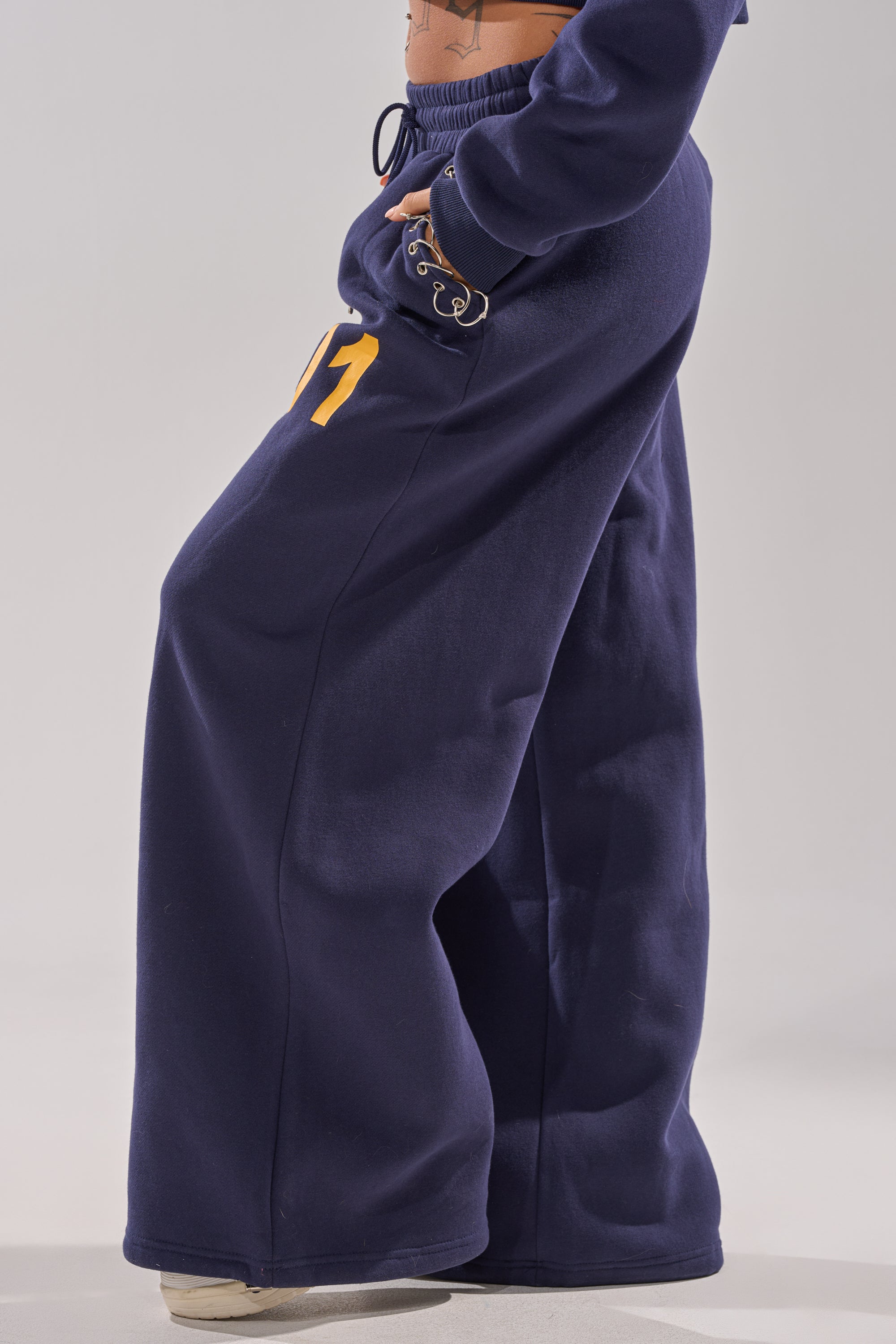 Someone stands against a plain light background wearing the UNTAMED METAL RING JOGGER—wide-leg fleece sweatpants in navy blue with yellow numbers, a drawstring waistband—and white shoes. 3