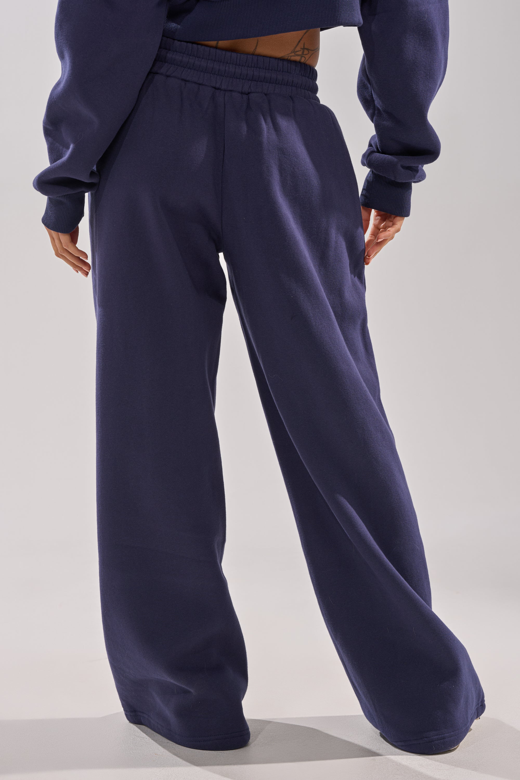 A person wearing the UNTAMED METAL RING JOGGER—a pair of loose-fitting navy blue wide-leg sweatpants—pose with their back to the camera against a plain light background, paired with a matching sweatshirt. 4