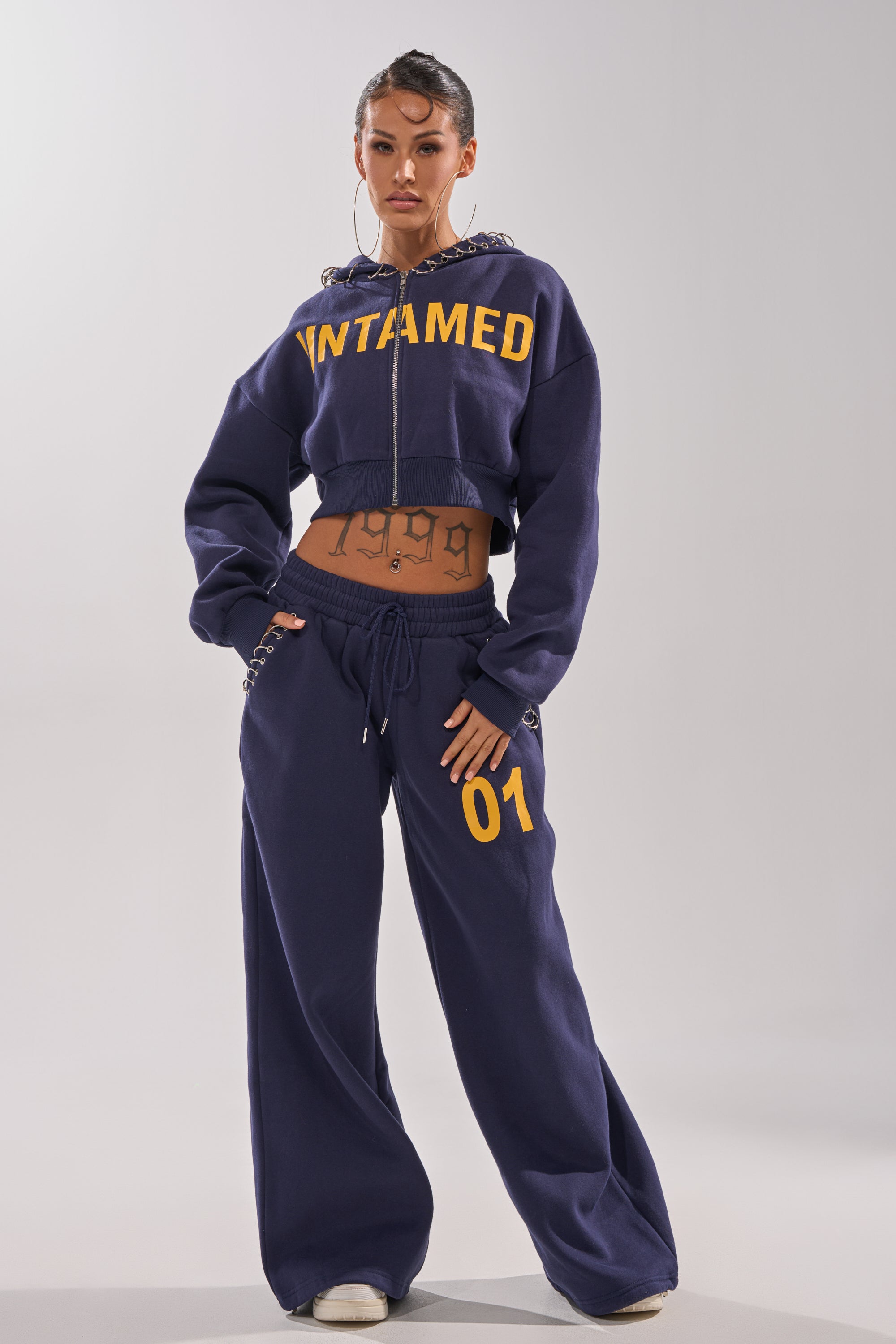 A woman poses confidently in the UNTAMED METAL RING JOGGER set—navy blue with "UNTAMED" in yellow and "01." details—showing tattoos on her torso and wearing large hoop earrings. 5