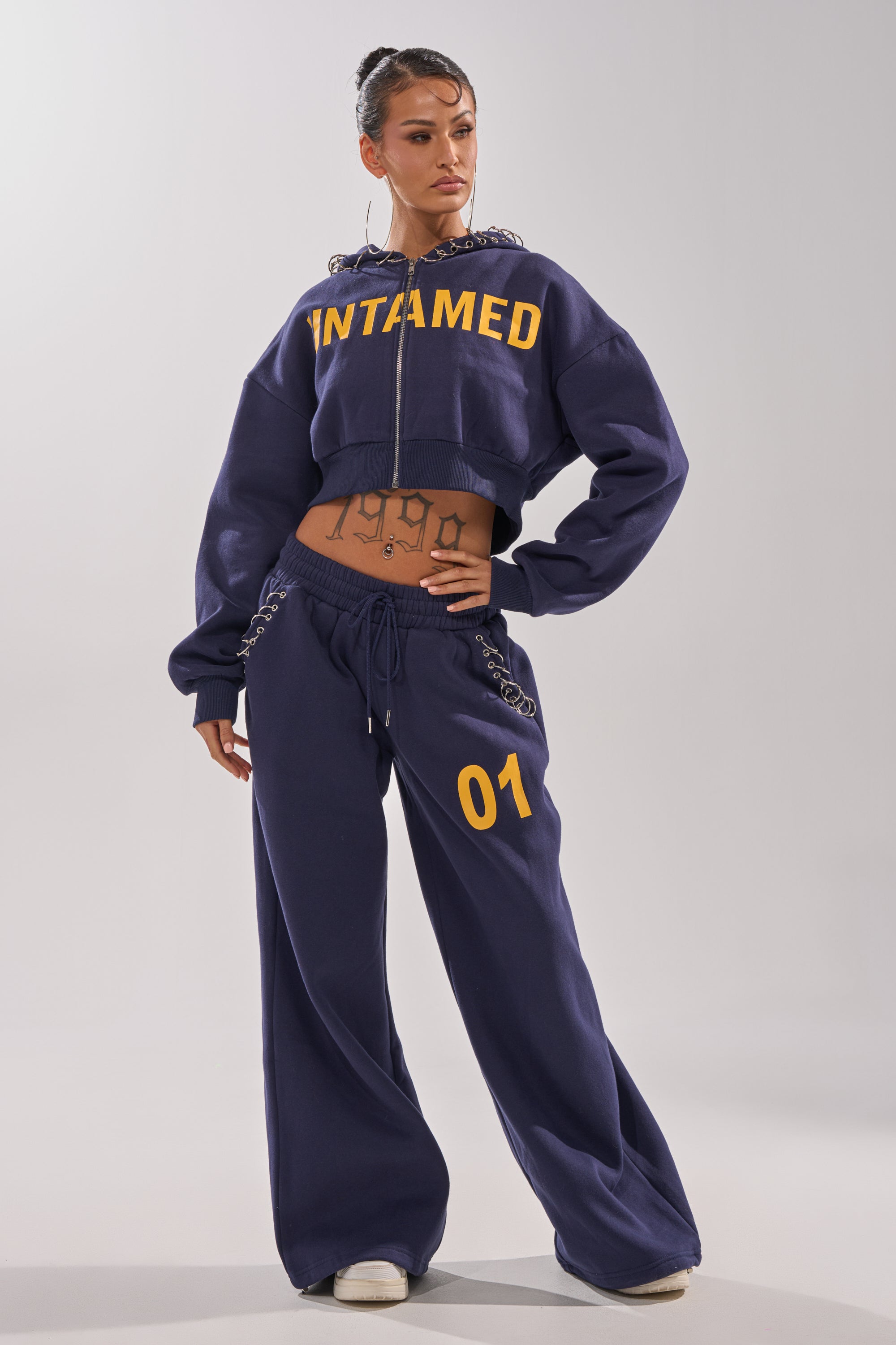 A woman models the UNTAMED METAL RING JOGGER, featuring "UNTAMED" on the cropped jacket and "01" on the high-rise pants, revealing her stomach tattoos against a simple light background. 6