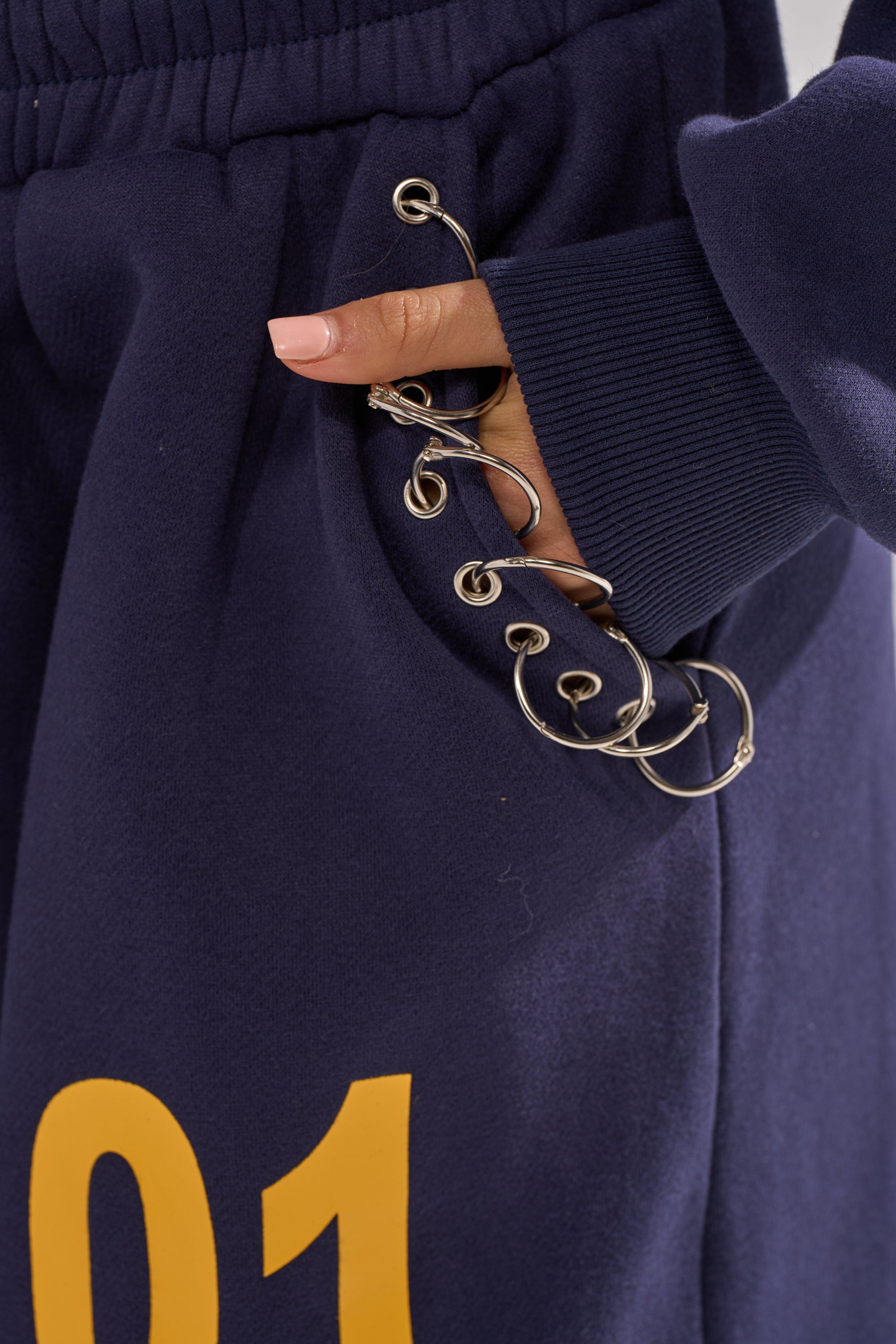 A close-up of a hand in the pocket of the UNTAMED METAL RING JOGGER, navy fleece sweatpants featuring a yellow "01" and metallic rings decorating the pocket opening for a distinctive look. 8