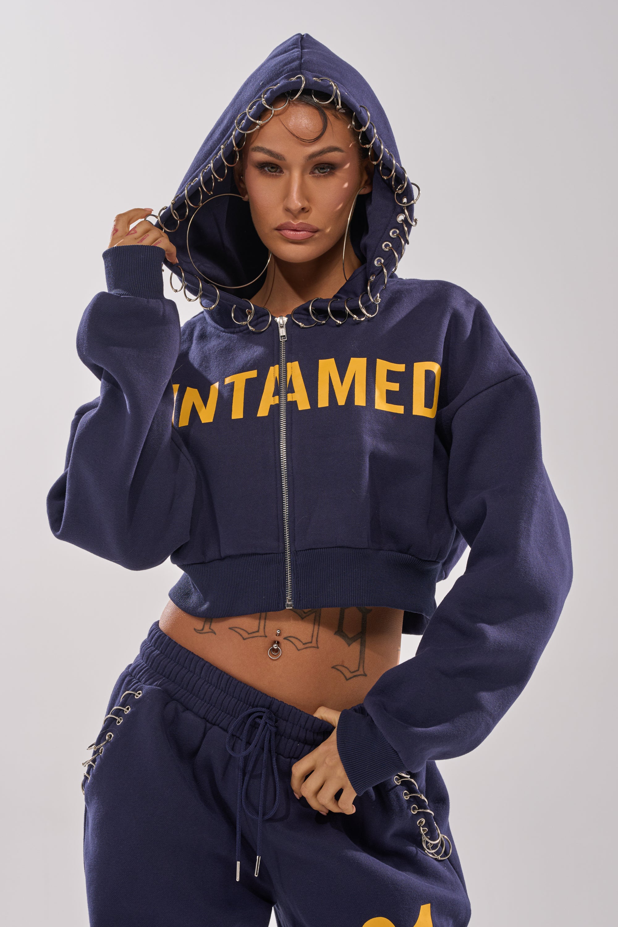 A woman models the UNTAMED METAL RING SWEATSHIRT in navy with yellow lettering and metal rings on the hood's edge, paired with matching sweatpants. She holds the hood and looks confidently at the camera.