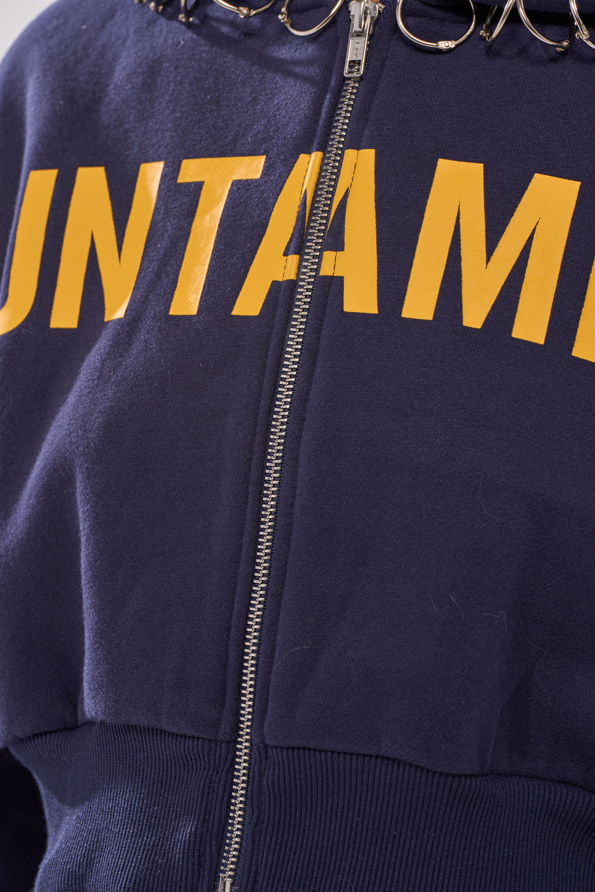 Close-up of the UNTAMED METAL RING SWEATSHIRT, a navy cropped zip-up hoodie with bold yellow "UNTAAM" lettering and metal rings at the collar for a striking women's street style look.