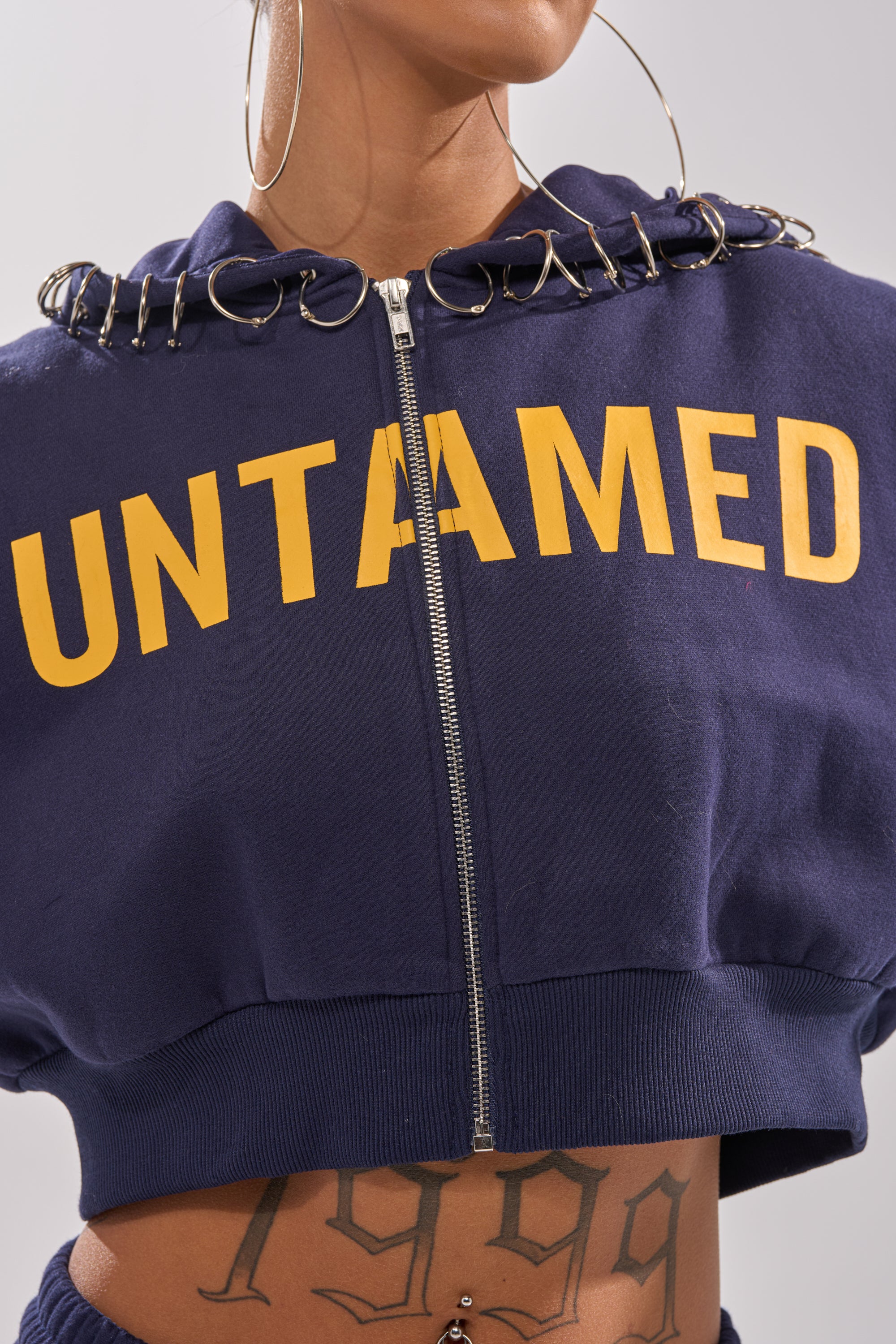 Wearing the UNTAMED METAL RING SWEATSHIRT with bold yellow graphics, metal ring accents on the hood, large hoop earrings, a "1999" stomach tattoo, and a belly button piercing for a standout women’s street style look.