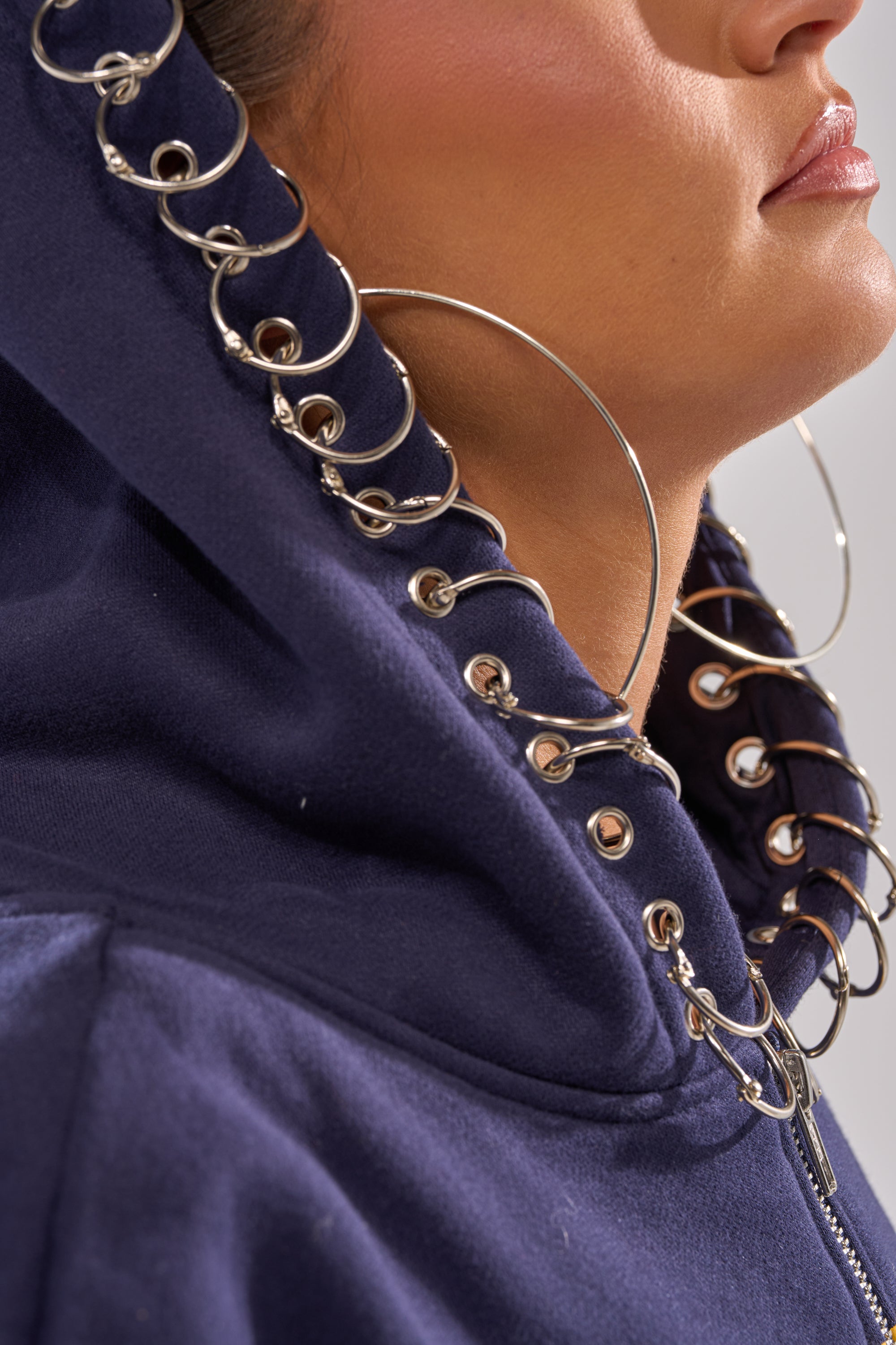 A close-up of a person wearing the UNTAMED METAL RING SWEATSHIRT, featuring silver rings through the hood and large hoop earrings. Their partially visible face highlights bold women’s street style with focus on jawline and lips.