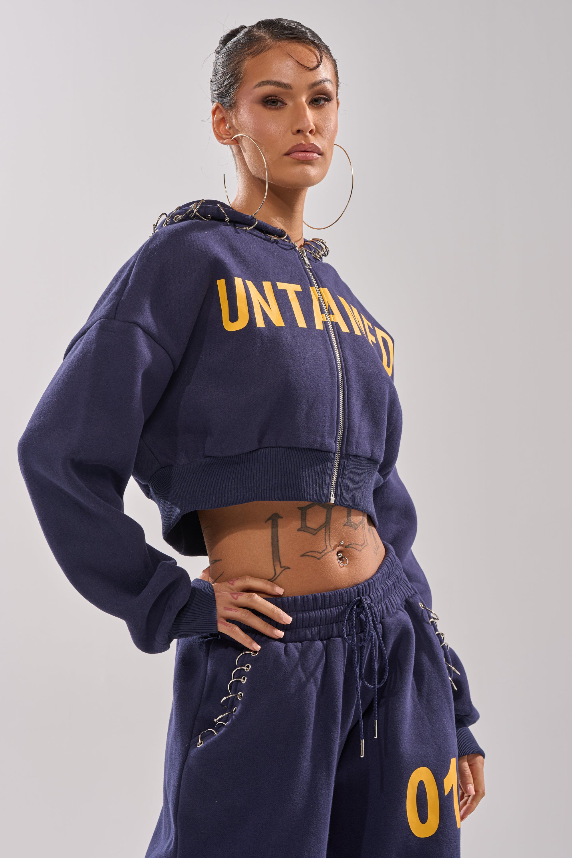 A woman stands confidently in the UNTAMED METAL RING SWEATSHIRT, paired with matching navy joggers marked "07," large hoop earrings, and visible tattoos on her torso and hand.