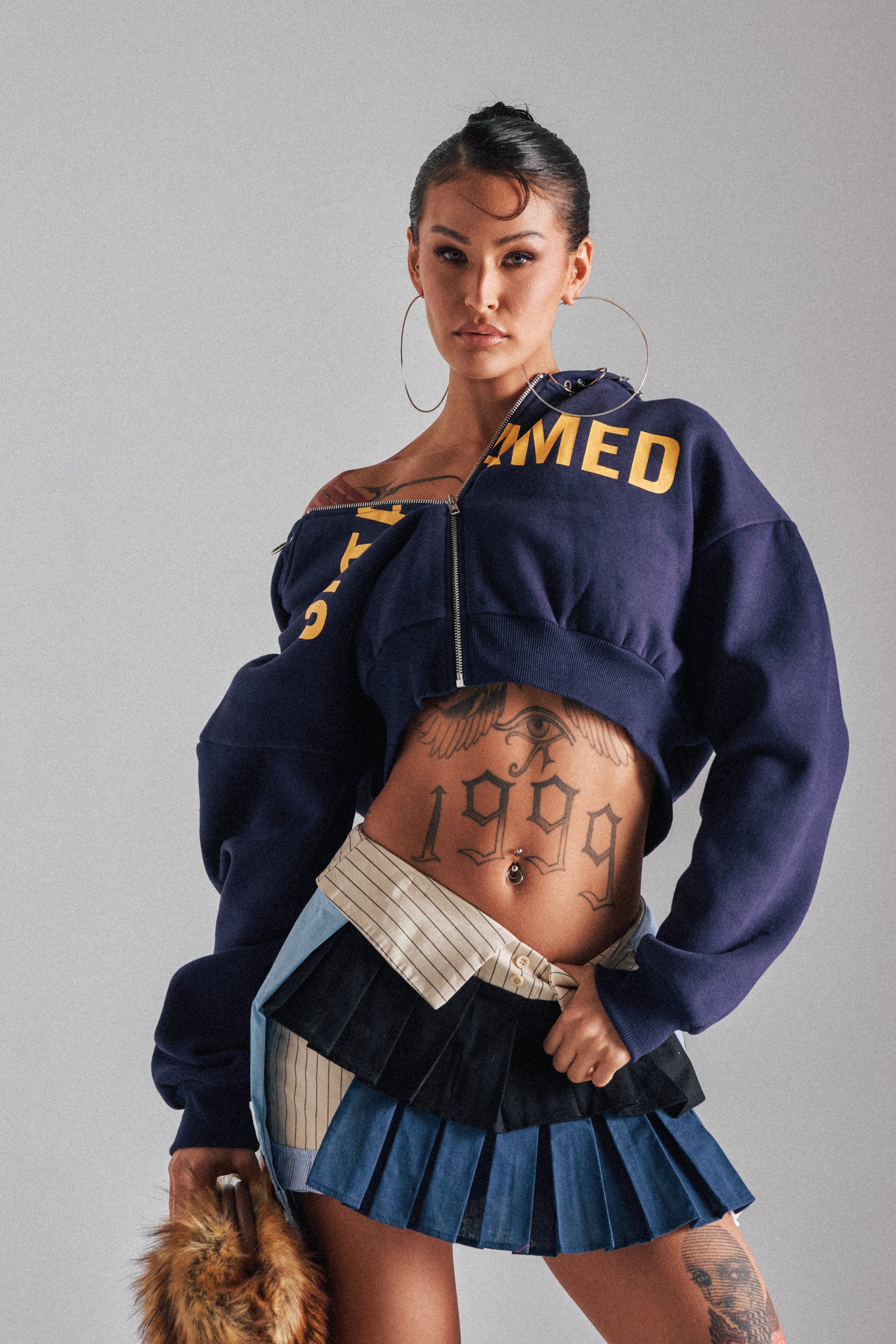A confident woman with visible tattoos, including "1999" on her stomach, wears the UNTAMED METAL RING SWEATSHIRT styled with hoop earrings, a pleated mini skirt, and holds a small furry bag.