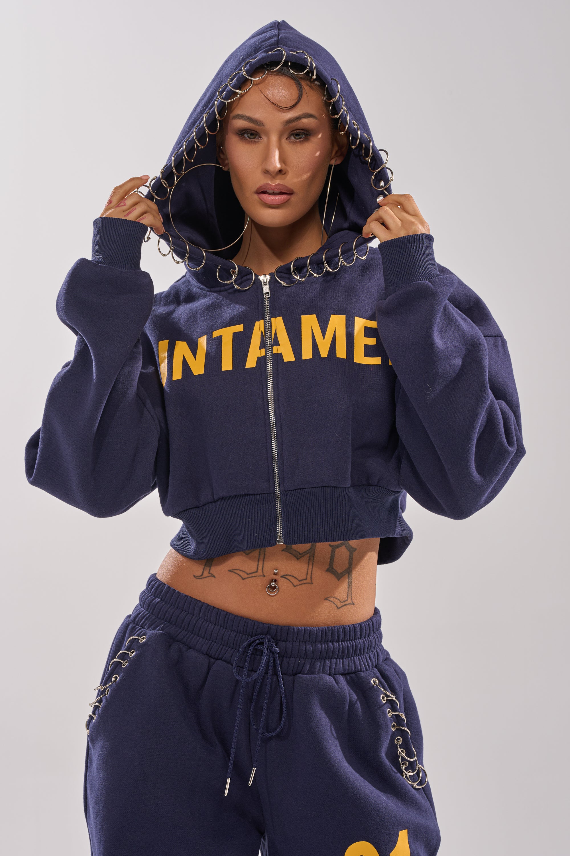 A person poses street style in the UNTAMED METAL RING SWEATSHIRT, a navy cropped zip-up hoodie with "UNTAMED" in yellow and metal ring details, paired with matching sweatpants and the hood up.