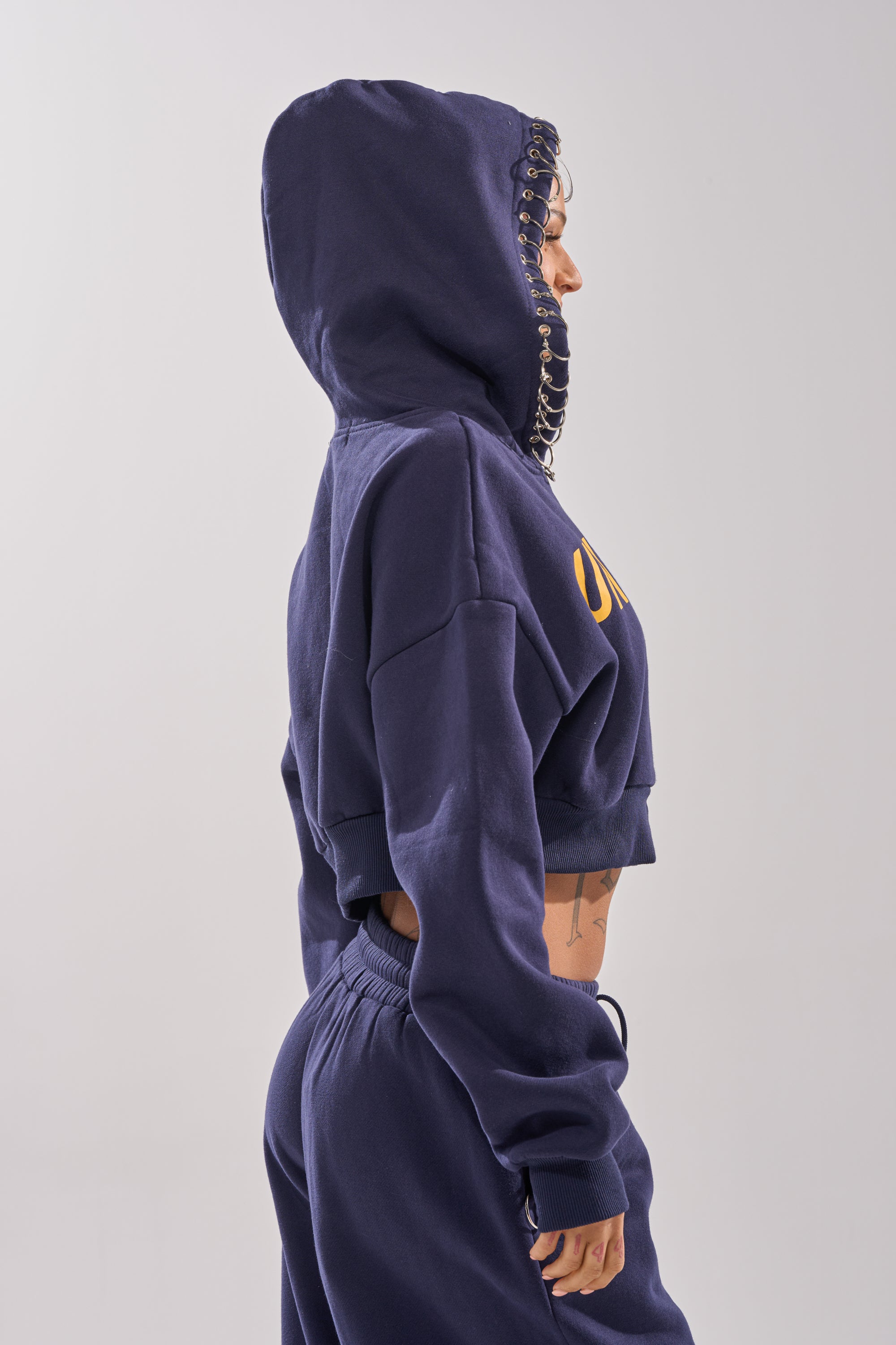 Wearing the UNTAMED METAL RING SWEATSHIRT and matching joggers, a person stands in profile against a plain background. The cropped, navy zip-up hoodie with metal hood rings reveals a torso tattoo, highlighting bold street style.