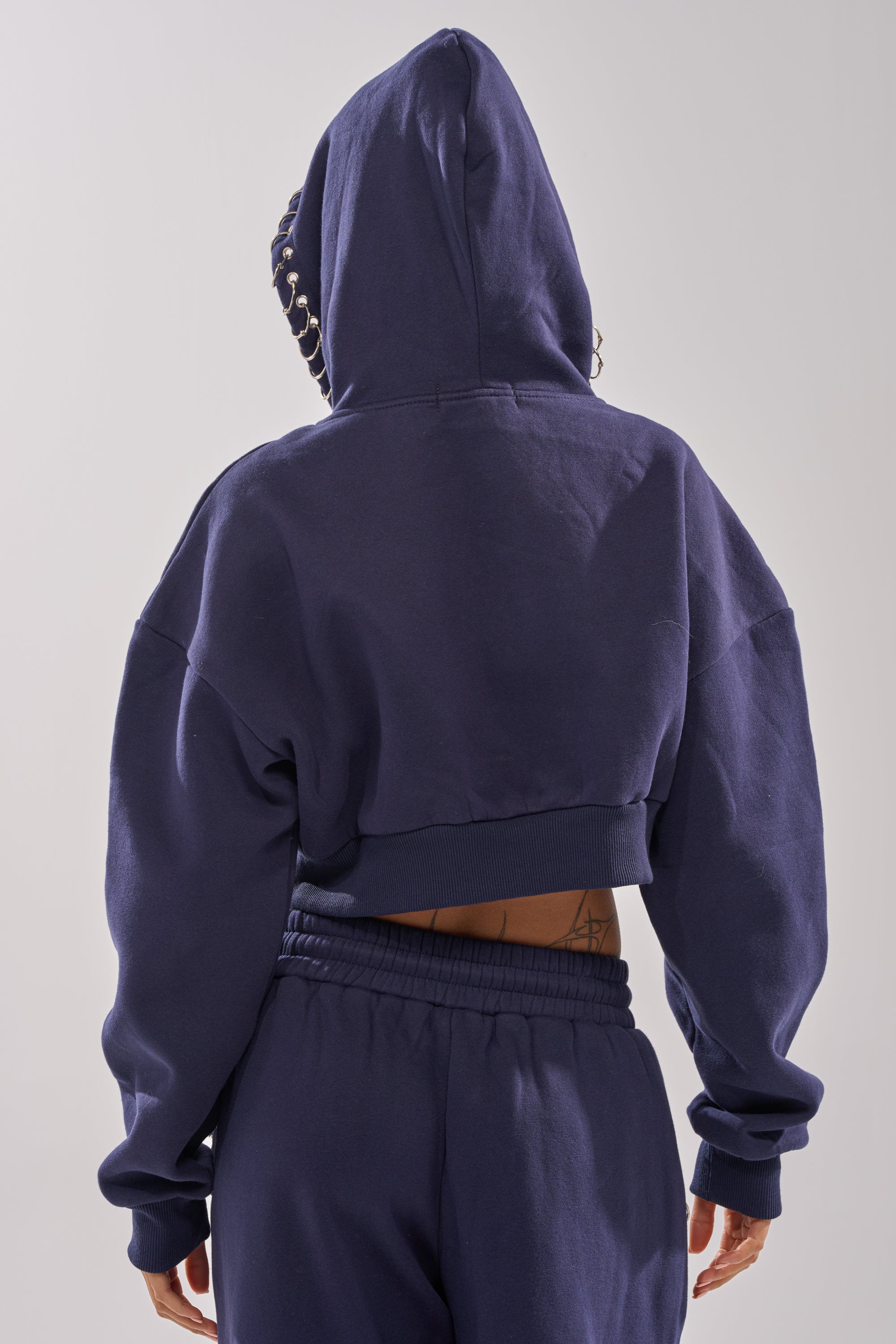 A person is shown from the back wearing the UNTAMED METAL RING SWEATSHIRT—a dark blue cropped zip-up hoodie with the hood up and hands by their sides—revealing part of their lower back for a trendy street style look.