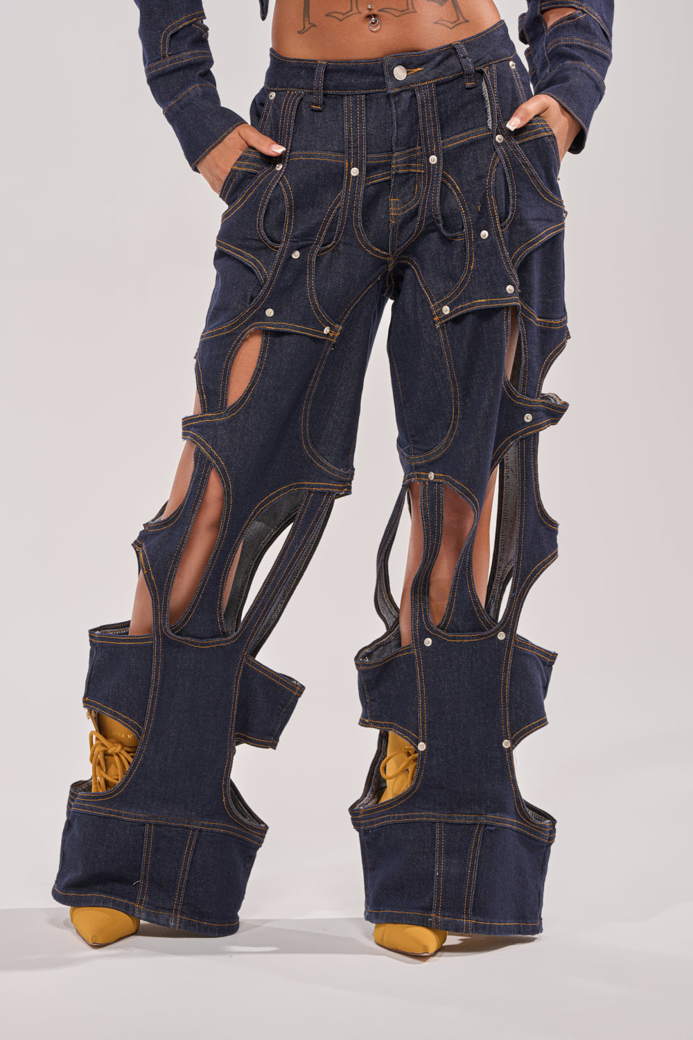 Wearing the CUT YOU OUT RELAXED DENIM—unique, deconstructed jeans with large cutouts and visible brown stitching that reveal skin and show yellow shoes through the open ankle sections. 0