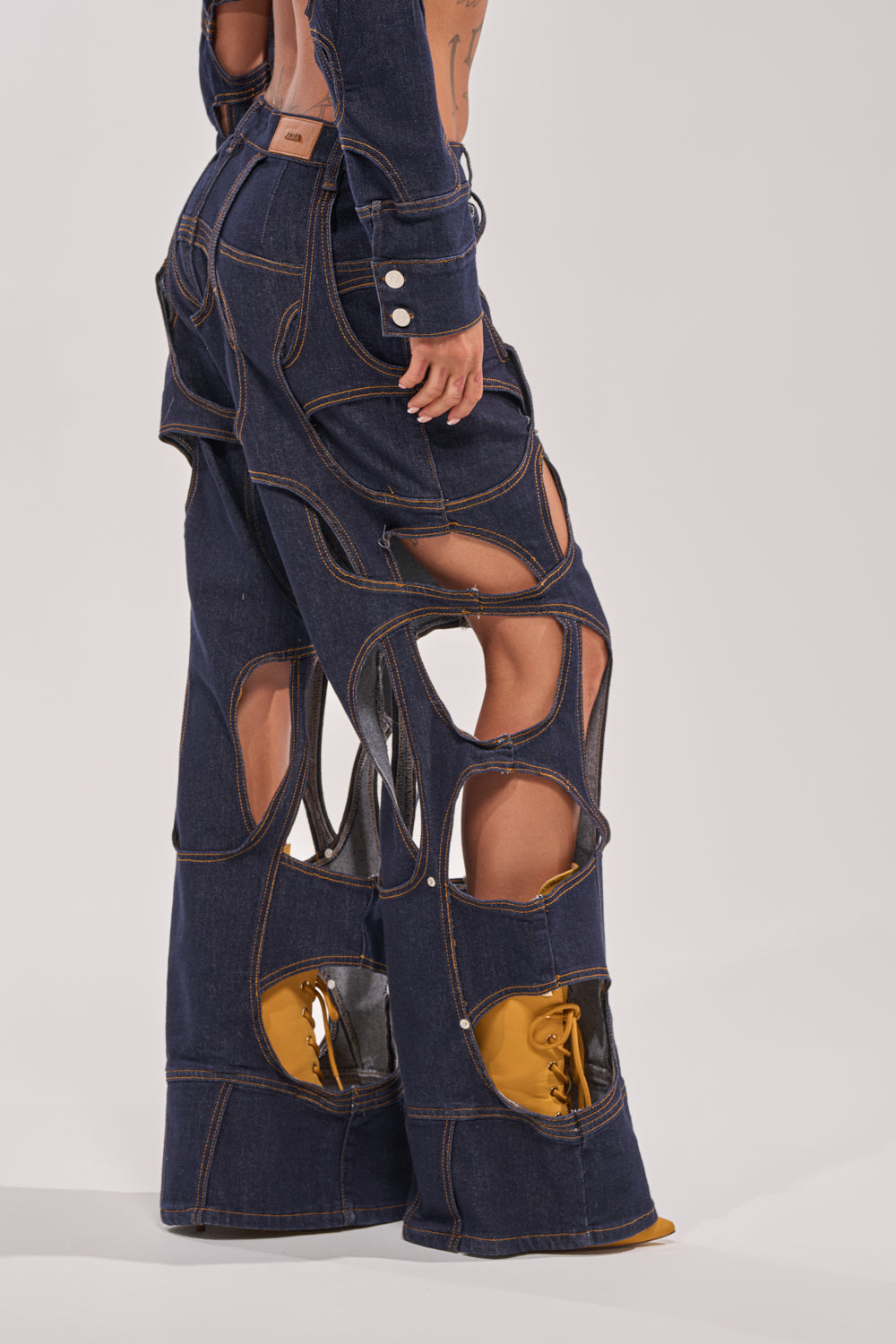 Wearing CUT YOU OUT RELAXED DENIM with bold, large cutouts and visible seams, paired with yellow shoes, this look stands out against a plain light background, creating an avant-garde fashion statement. 1