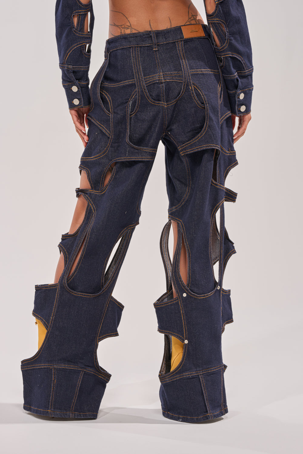 A person viewed from the back wears CUT YOU OUT RELAXED DENIM: dark blue deconstructed jeans with large cutouts revealing yellow fabric, exaggerated seams, and wide structured legs for a bold statement. 2