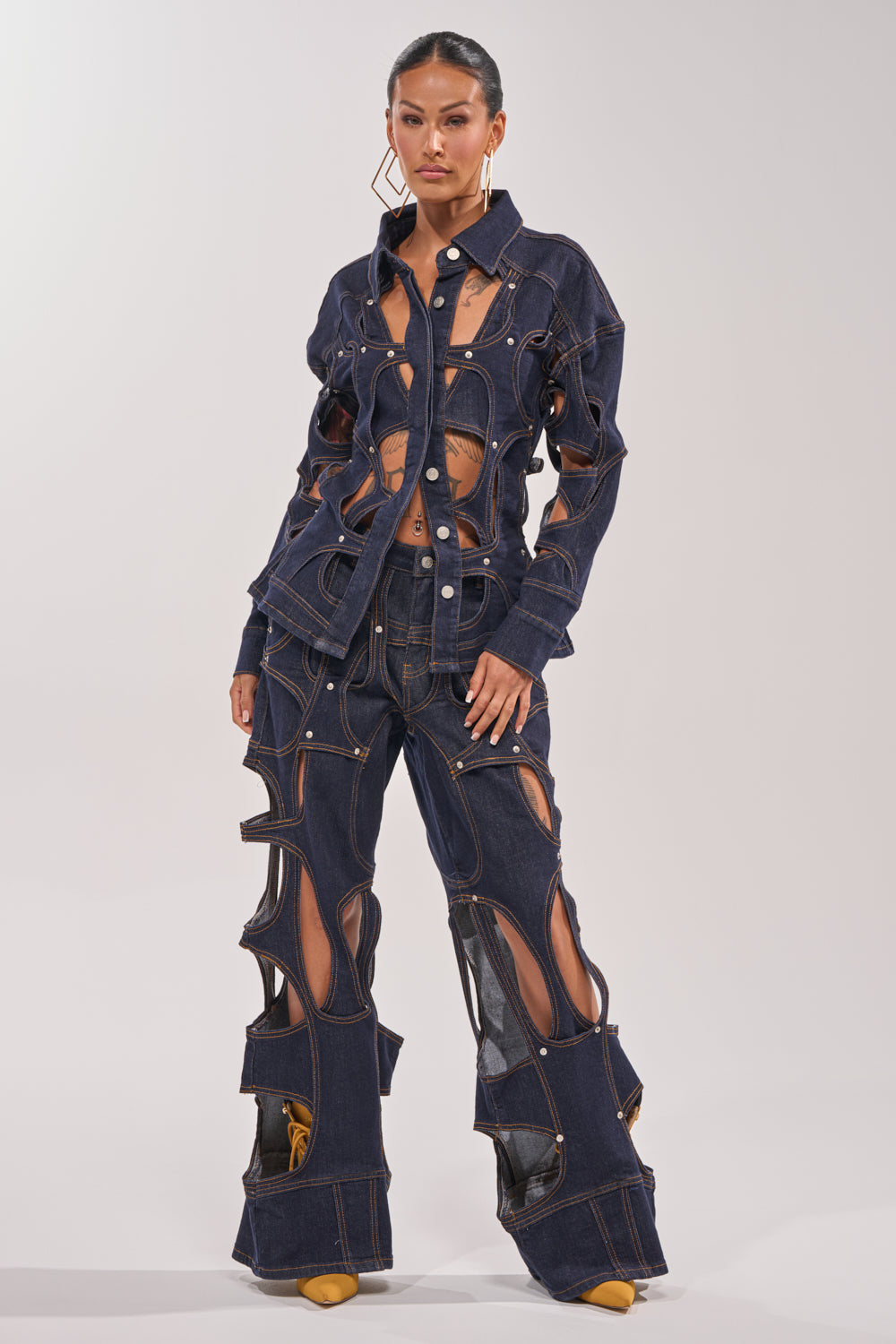 Wearing the CUT YOU OUT RELAXED DENIM, a woman poses in a unique deconstructed jeans outfit with large cutouts on the jacket and flared pants, styled with geometric gold earrings and yellow shoes against a plain light background. 3