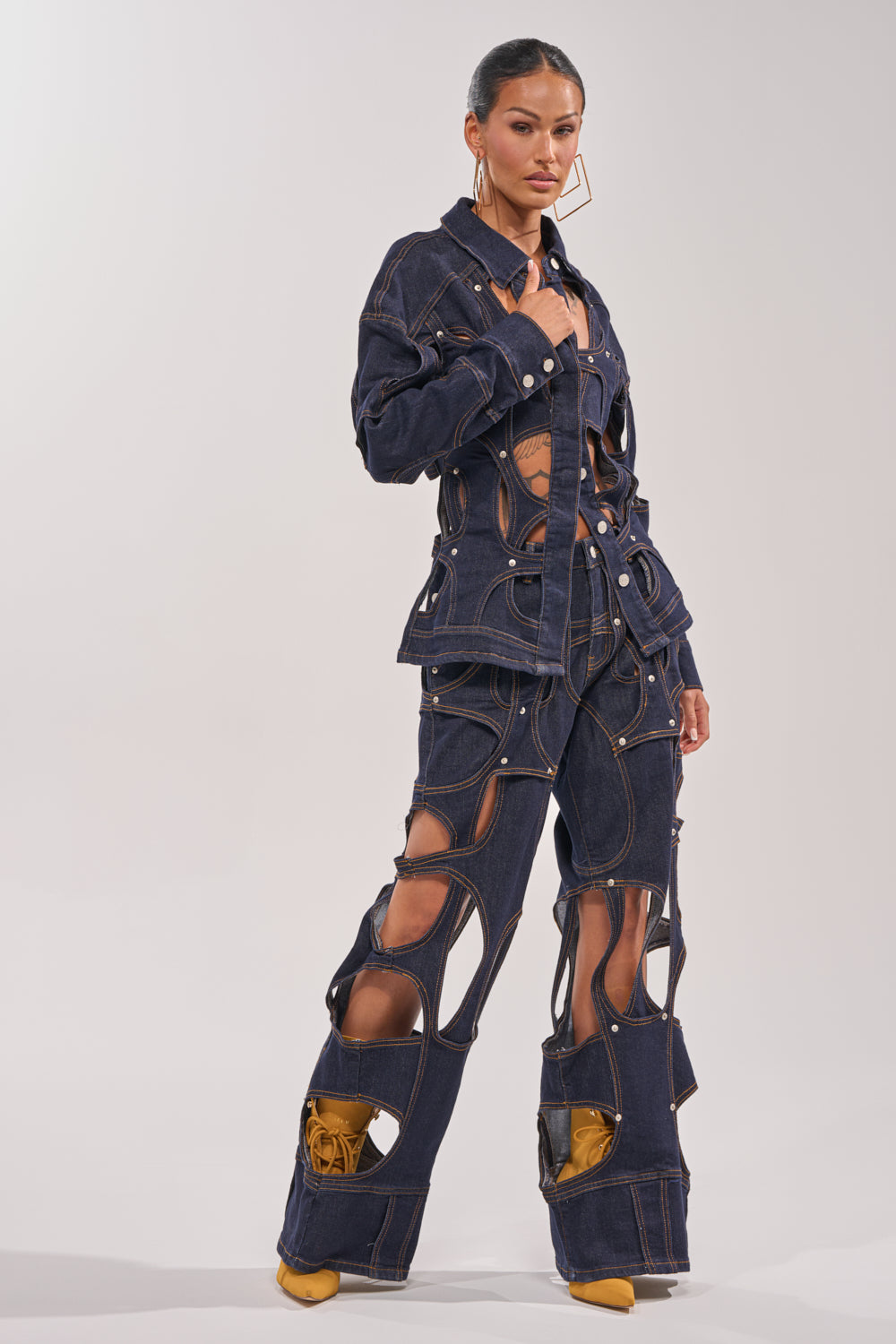 A woman models the CUT YOU OUT RELAXED DENIM, featuring bold cutouts on the jacket and straight-leg pants that reveal skin and a torso tattoo. She accessorizes with gold hoop earrings and yellow heels, standing against a simple backdrop. 4