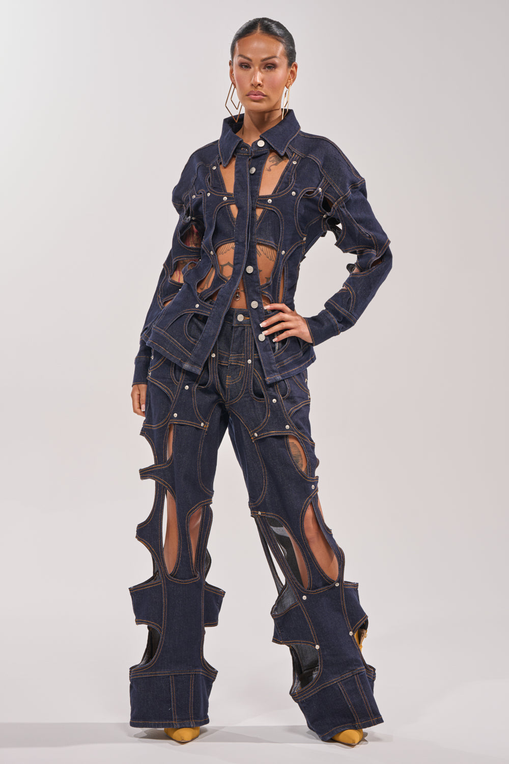 Wearing the CUT YOU OUT RELAXED DENIM, a person poses confidently in a dark wash straight-leg set featuring bold cutouts on the jacket and pants, accented by yellow shoes and large hoop earrings against a simple light background. 5