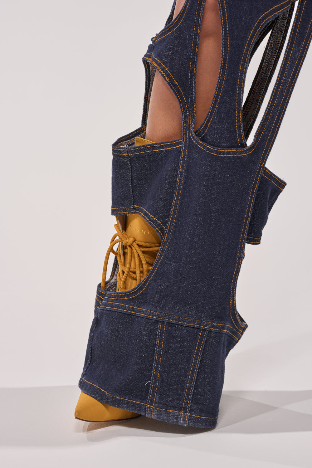 A close-up of a foot in a yellow pointed-toe shoe with laces, partially covered by CUT YOU OUT RELAXED DENIM featuring exposed seams and creative openings that reveal glimpses of the leg and shoe. 6