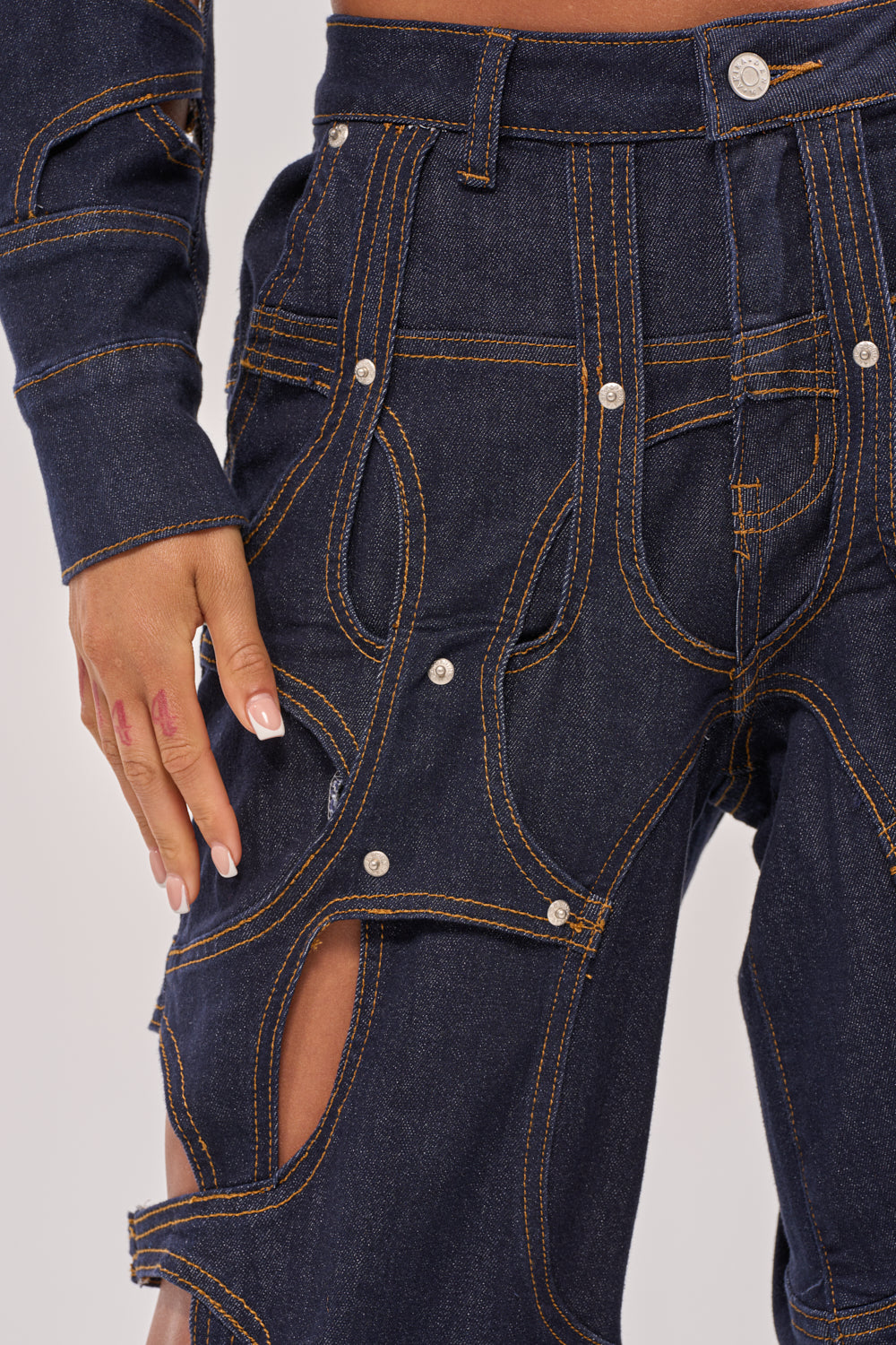 Close-up of a person wearing CUT YOU OUT RELAXED DENIM pants featuring unique cutout designs and exposed contrasting stitching, paired with a matching denim jacket sleeve. Their hand rests near the hip. 7