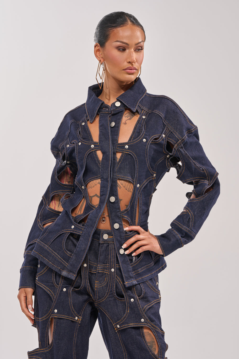 Wearing the CUT YOU OUT BUTTON DOWN with bold cutout details, a woman stands confidently with one hand on her hip, tattoos visible beneath the deconstructed denim against a plain background. 0