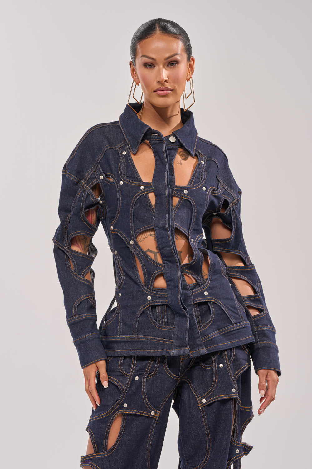 A woman models the CUT YOU OUT BUTTON DOWN in dark denim with cutout and circular details, paired with matching pants. She stands confidently against a plain background, hair pulled back and geometric earrings finishing her look. 1