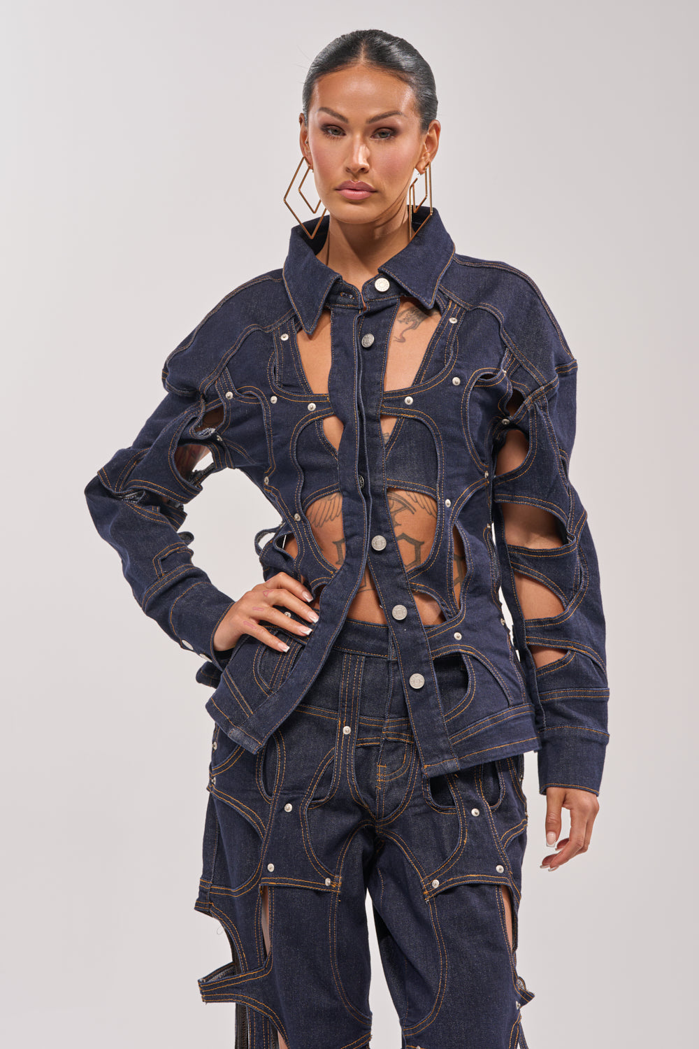 A woman models the CUT YOU OUT BUTTON DOWN, a dark denim jacket and pants set with cutout details and exposed skin. She pairs it with large geometric earrings, her hair slicked back, against a plain light background. 2