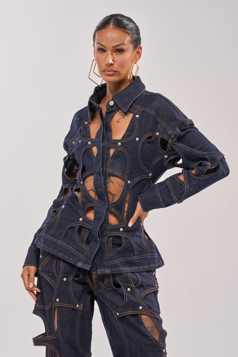 A woman models the CUT YOU OUT BUTTON DOWN, a dark denim piece featuring bold cutout details and exposed seams. She sports slicked-back hair, statement earrings, and a confident look against a plain light background. 3