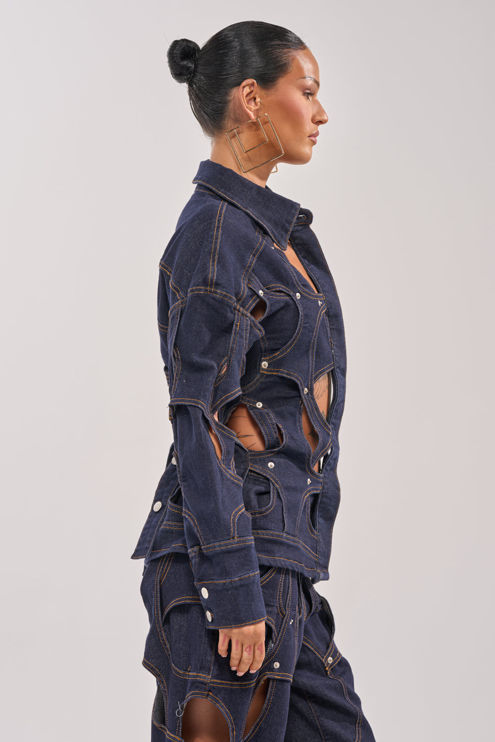 A woman with dark hair in a sleek bun stands in profile against a plain light background, wearing the CUT YOU OUT BUTTON DOWN—a unique denim piece with large cutout details and exposed seams. 4