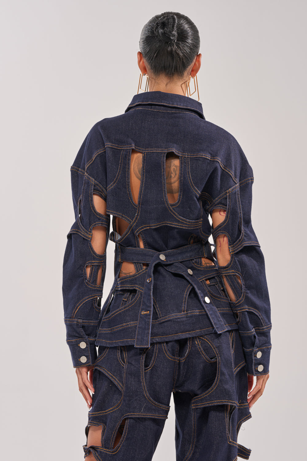 Seen from behind, a person with hair in a sleek bun wears the CUT YOU OUT BUTTON DOWN and matching pants in dark blue denim, featuring deconstructed silhouettes and distinctive cutout details that reveal patches of skin. 5