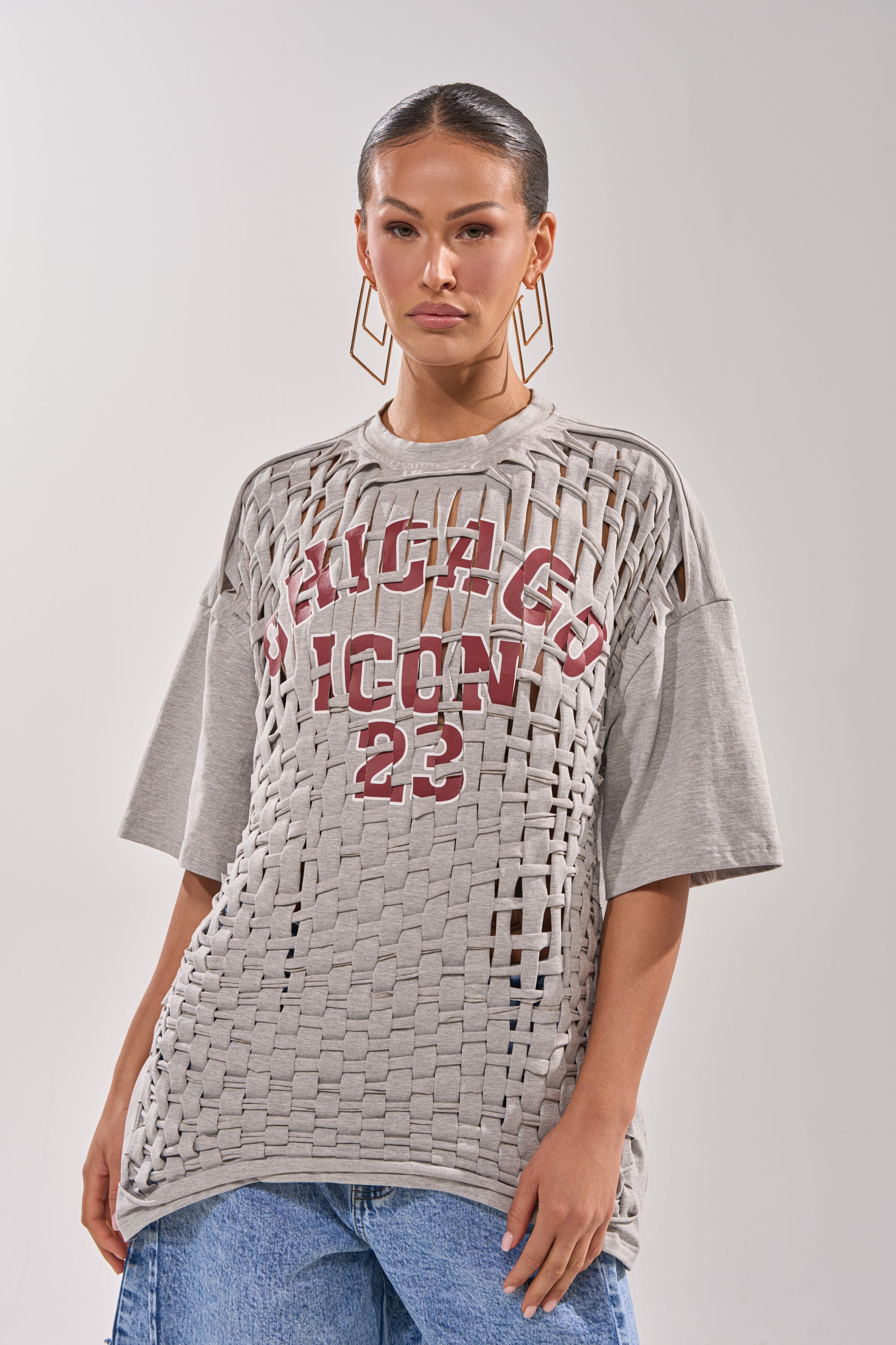 A woman wears the DISTRESSED FOR LIFE OVERSIZED TEE, a grey graphic shirt with Chicago lettering and "ICON 23" print, featuring a cutout overlay design. She pairs it with large hoop earrings and blue jeans against a plain background.