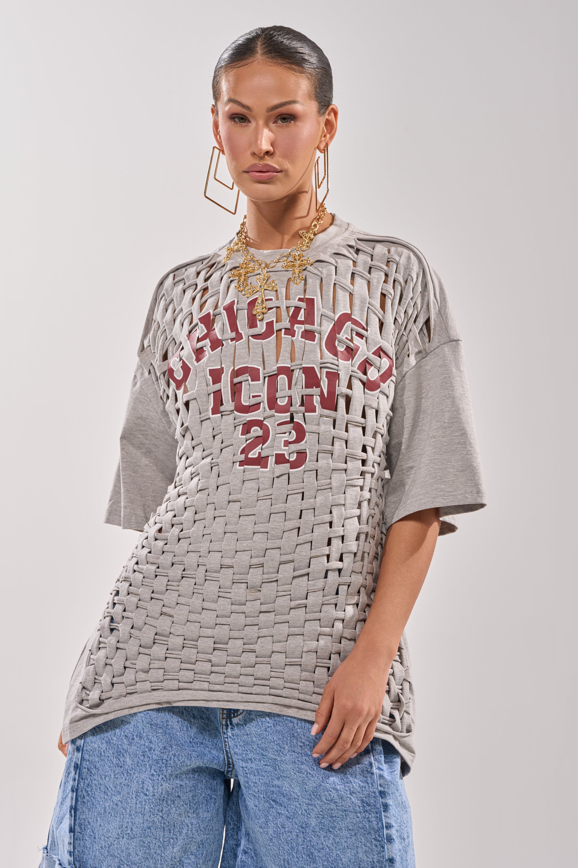 A woman models the DISTRESSED FOR LIFE OVERSIZED TEE—a grey graphic top with "Chicago Icon 23"—paired with large gold earrings, layered necklaces, and blue jeans, creating the ultimate festival look against a plain backdrop. 1