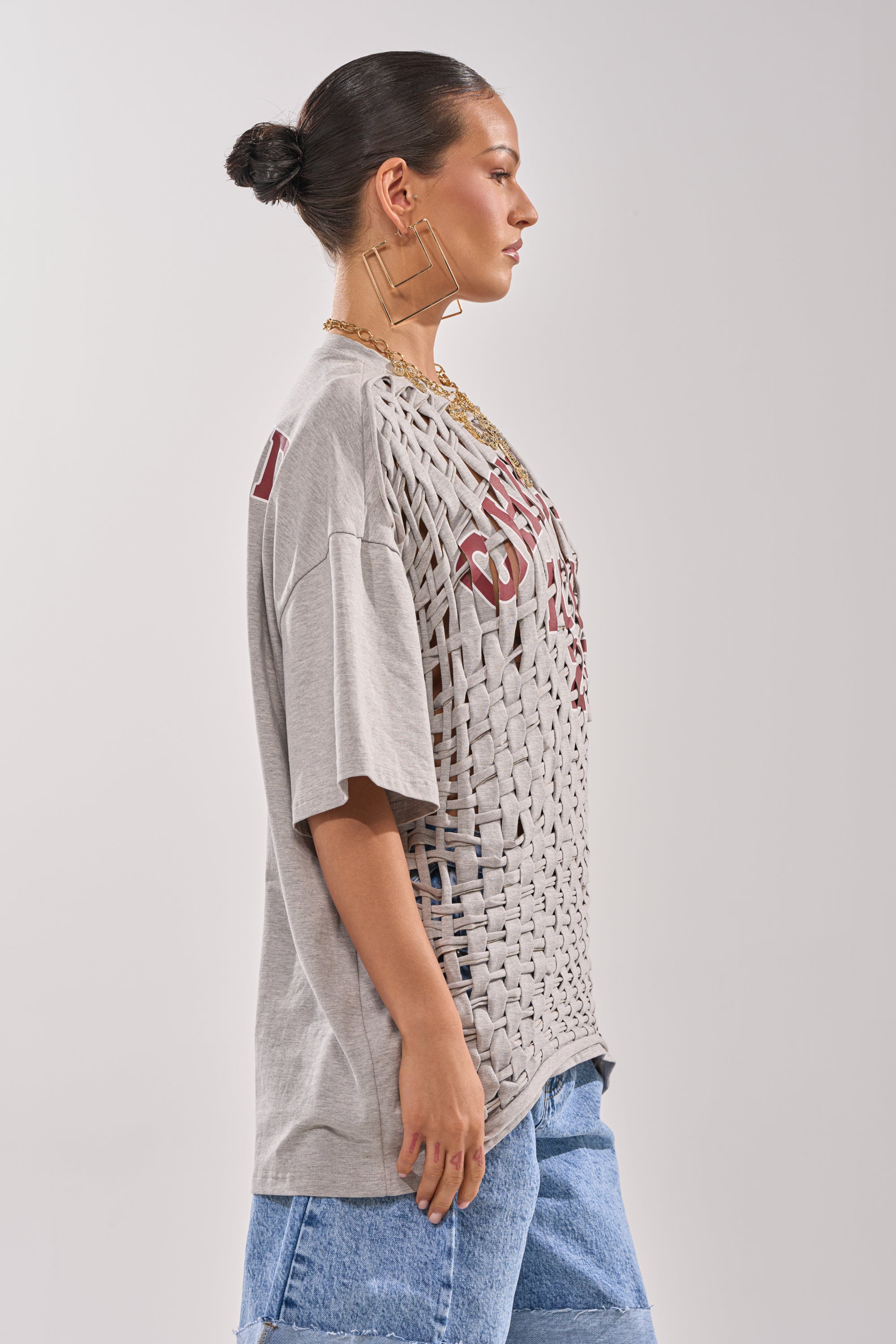 A woman in profile wears the DISTRESSED FOR LIFE OVERSIZED TEE, a loose grey graphic shirt, paired with blue denim shorts and large geometric earrings, her hair in a neat bun against a plain background. 4