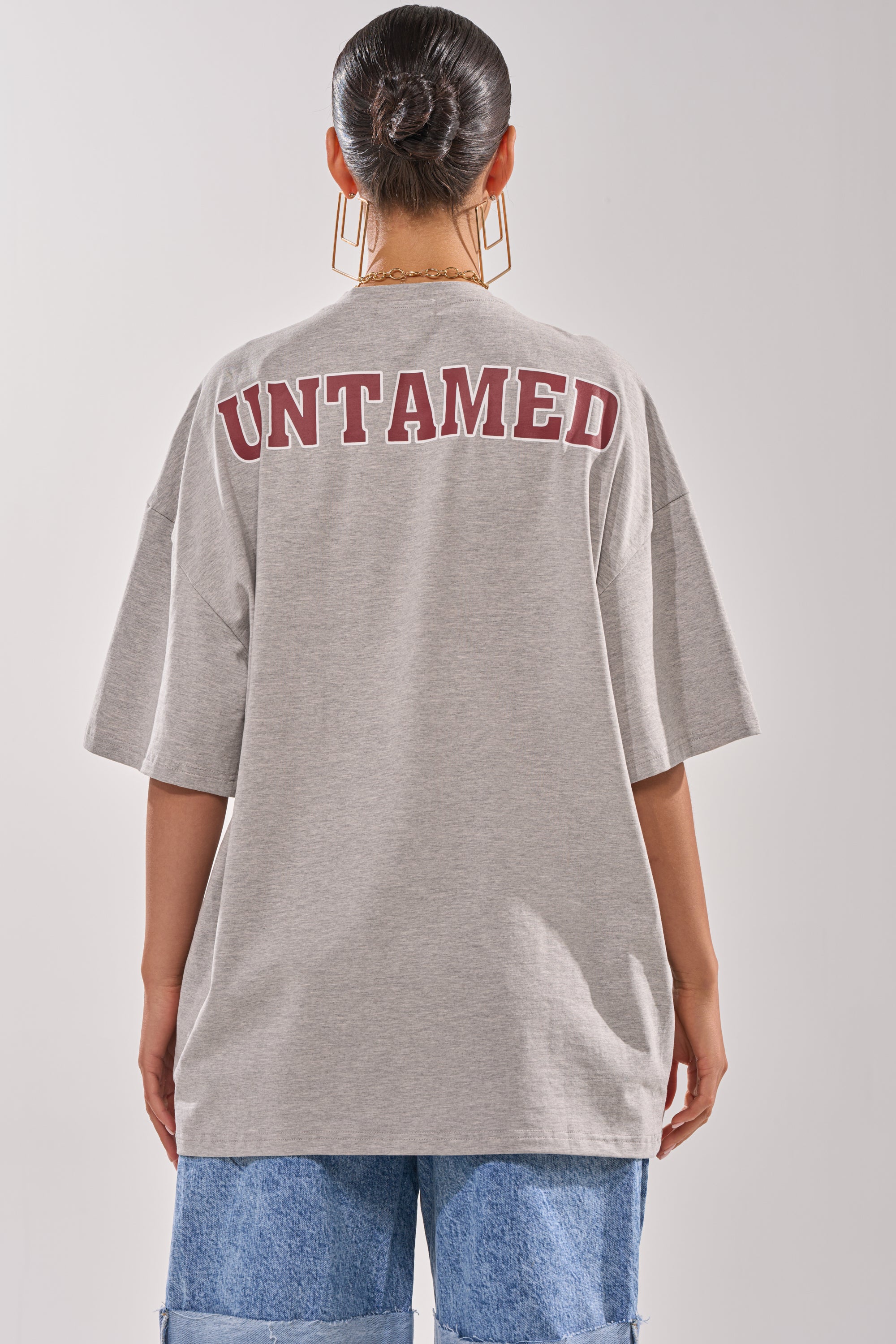 A person with dark hair in a bun wears the DISTRESSED FOR LIFE OVERSIZED TEE—a grey graphic shirt featuring "UNTAMED" in bold maroon letters on the back—paired with blue jeans, shown from behind against a plain background. 5