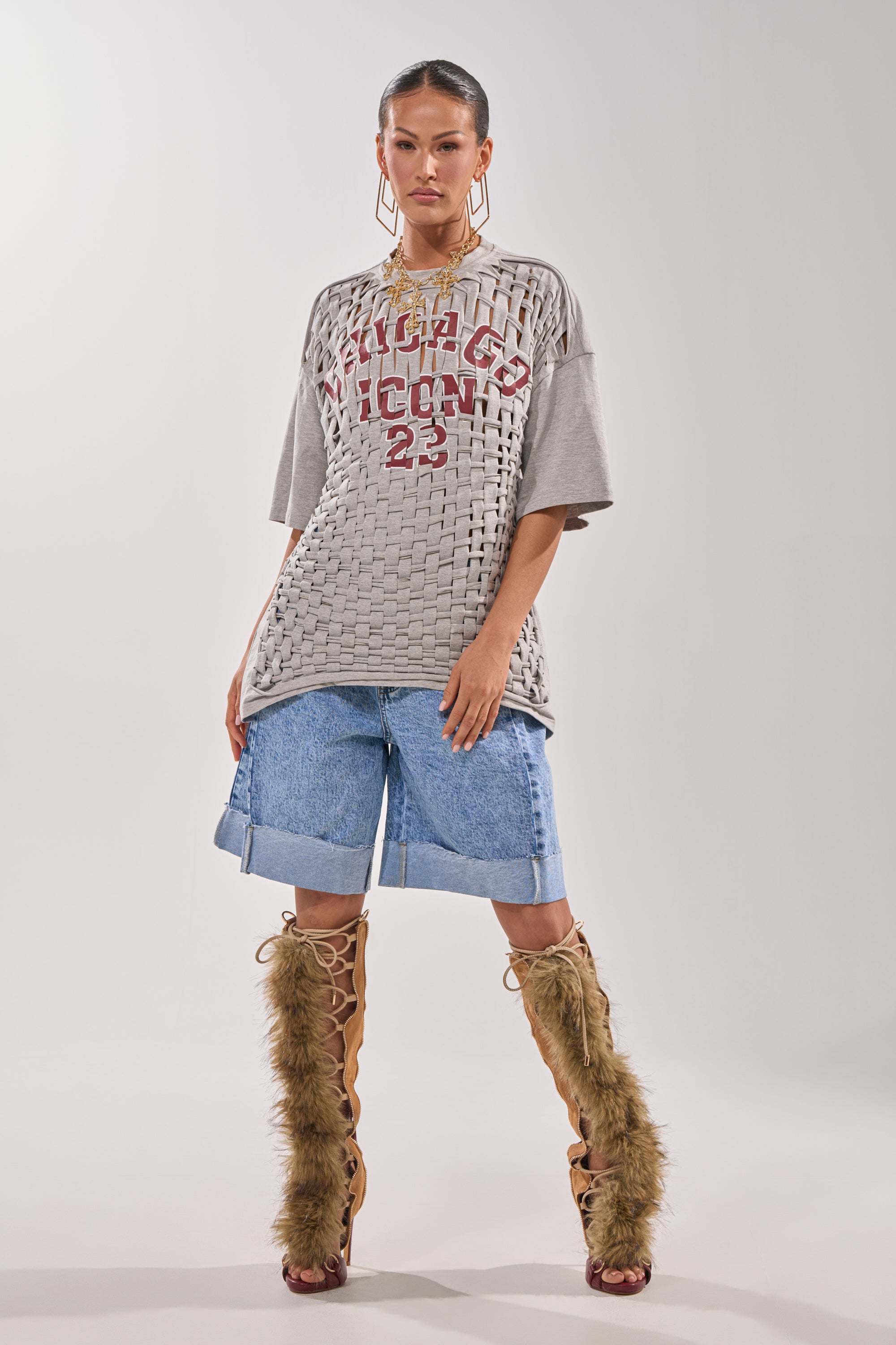 A woman stands against a plain background wearing the DISTRESSED FOR LIFE OVERSIZED TEE in grey, long denim shorts, knee-high lace-up boots with brown faux fur, large gold earrings, and a sleek pulled-back hairstyle. 6