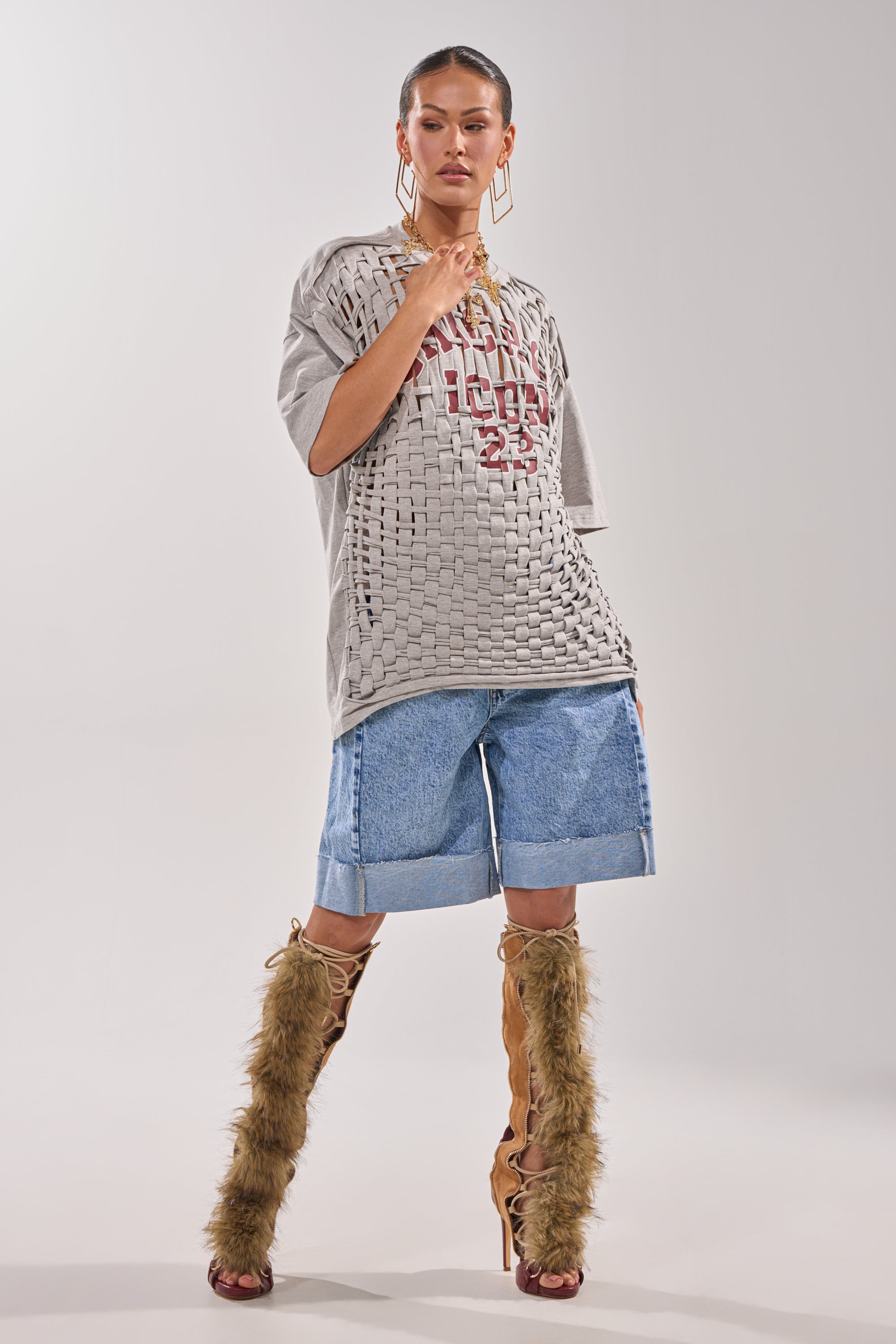 A woman poses confidently in the DISTRESSED FOR LIFE OVERSIZED TEE styled as a textured grey shirt with denim knee-length shorts, knee-high open-toe boots with brown fur, chunky gold jewelry, and slicked-back hair against a plain light background. 7
