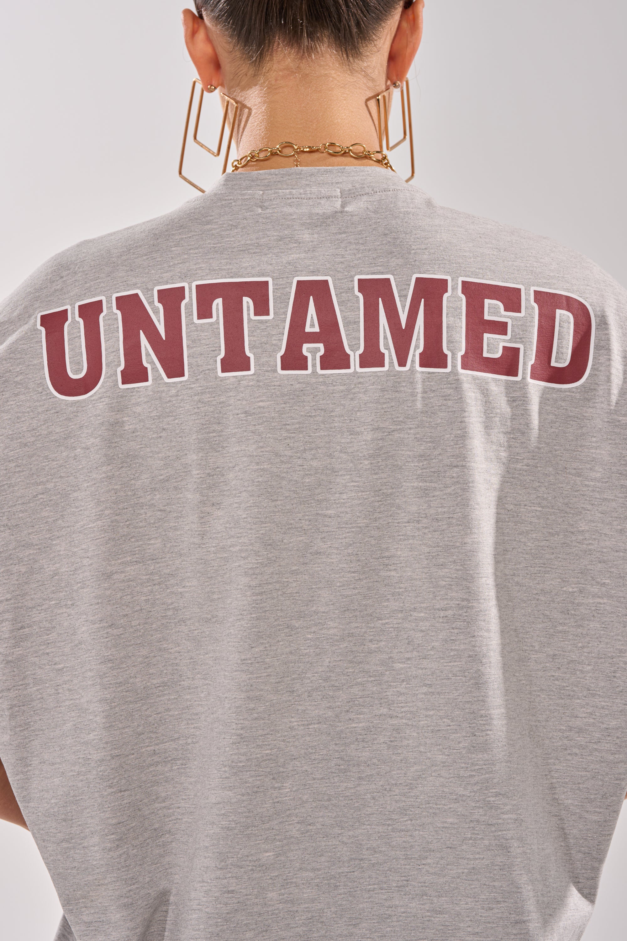 A person sports the DISTRESSED FOR LIFE OVERSIZED TEE, a grey graphic shirt with bold maroon "UNTAMED" lettering on the back, styled with short hair, chunky gold jewelry, and geometric earrings for an edgy urban vibe. 8