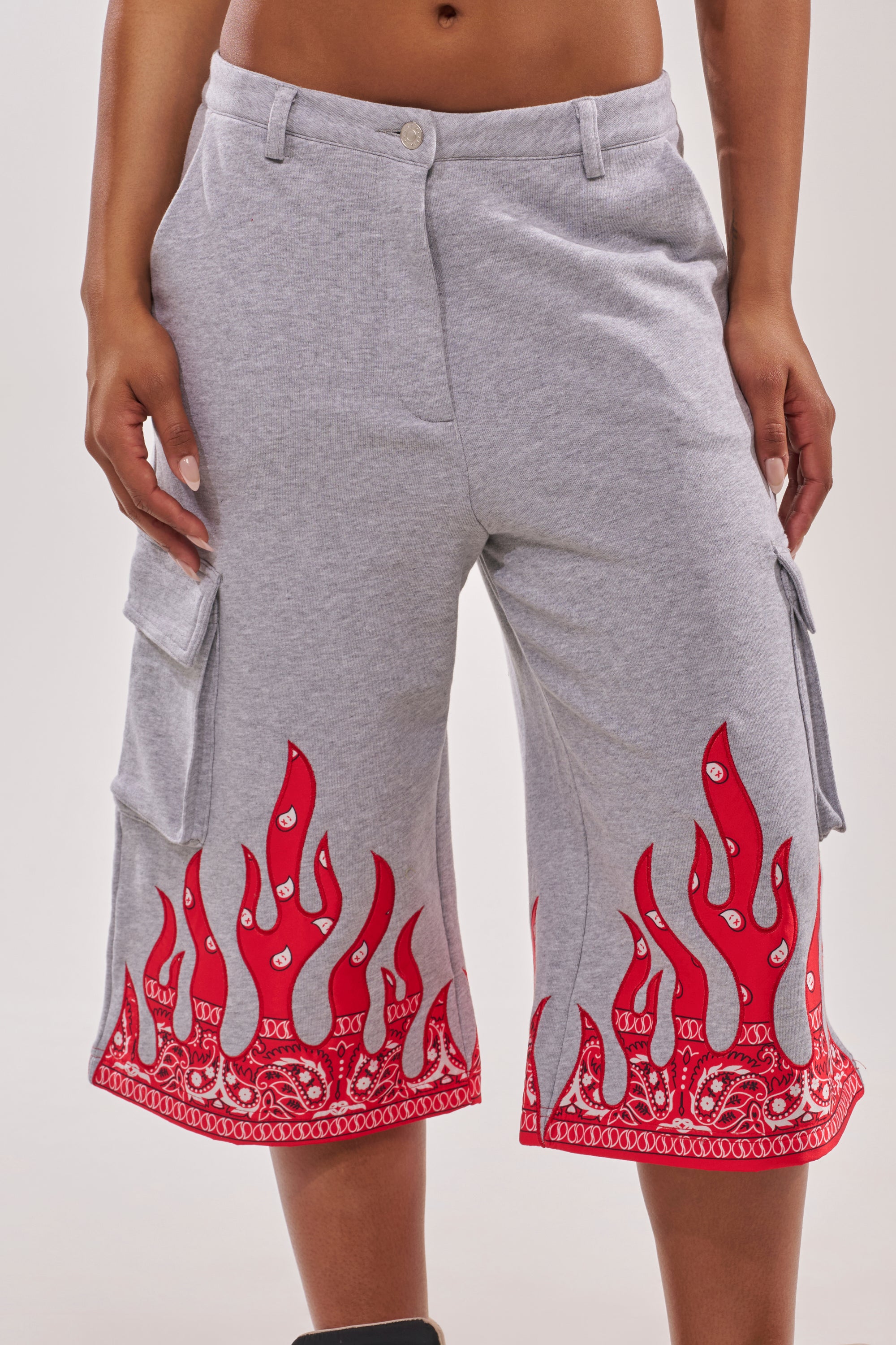 Someone stands against a plain background wearing the FIRE TO THE FLAMES BERMUDA SHORT, light gray cotton shorts with red flame and bandana patterns near the hem and cargo-style pockets. 1