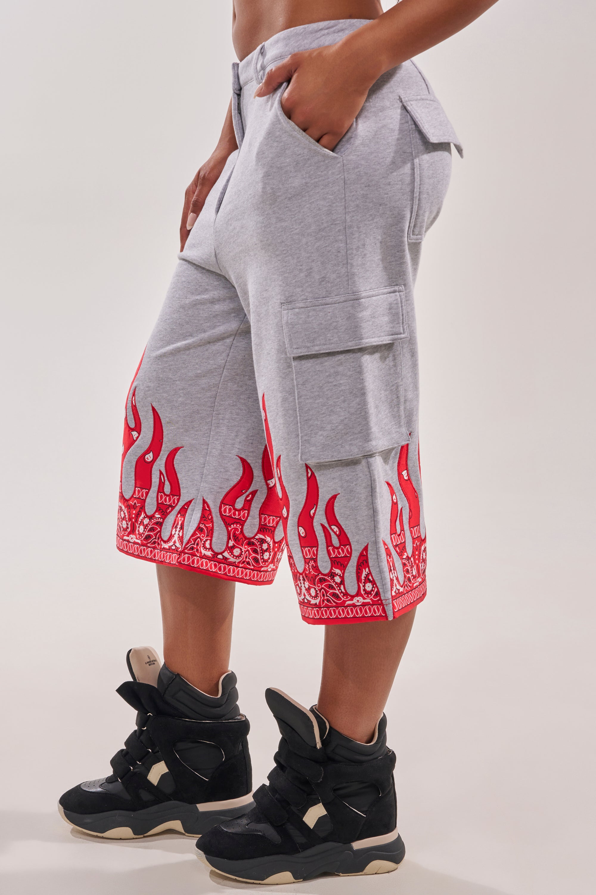 A person in FIRE TO THE FLAMES BERMUDA SHORT—light gray wide-leg shorts with red paisley flame hem detail—paired with black high-top wedge sneakers, stands sideways with hands in pockets. 3