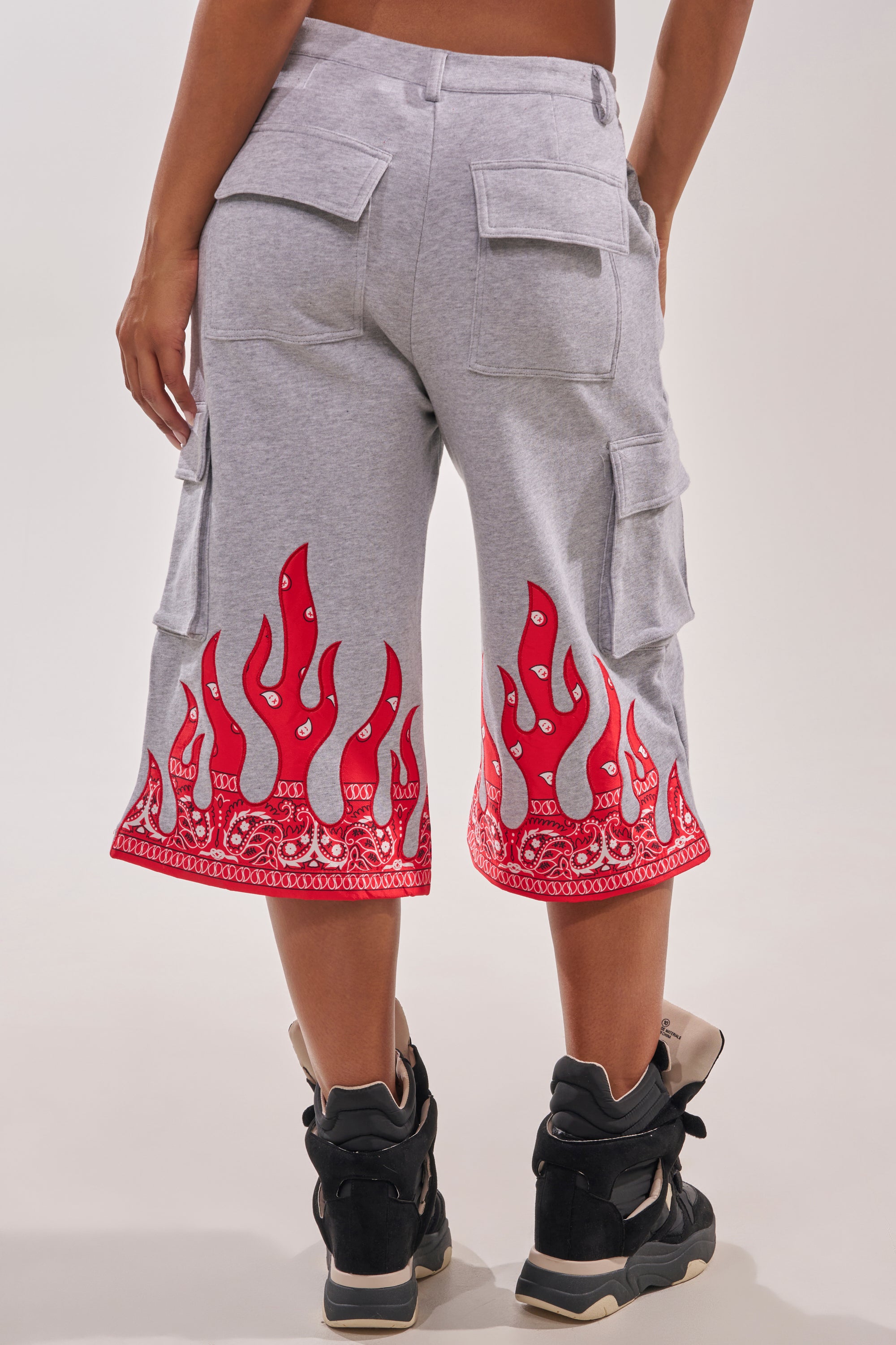 A person is shown from the back in FIRE TO THE FLAMES BERMUDA SHORT, featuring gray cotton fabric and red paisley flame details at the hem, paired with black and beige high-top sneakers against a plain background. 4