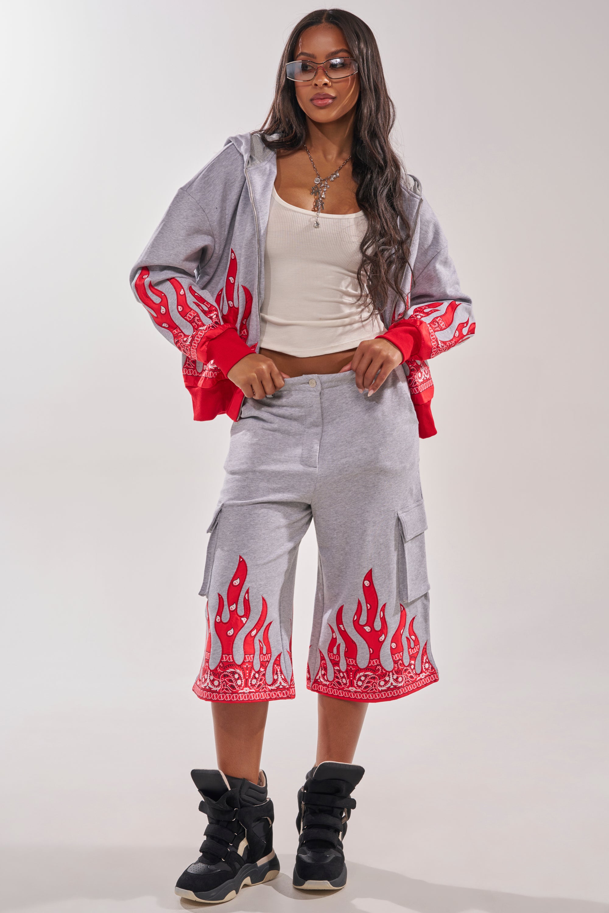 A woman stands confidently against a plain background wearing the FIRE TO THE FLAMES BERMUDA SHORT, featuring bold paisley flame detailing, paired with a gray hoodie, white tank top, black high-top sneakers, sunglasses, and layered necklaces. 6
